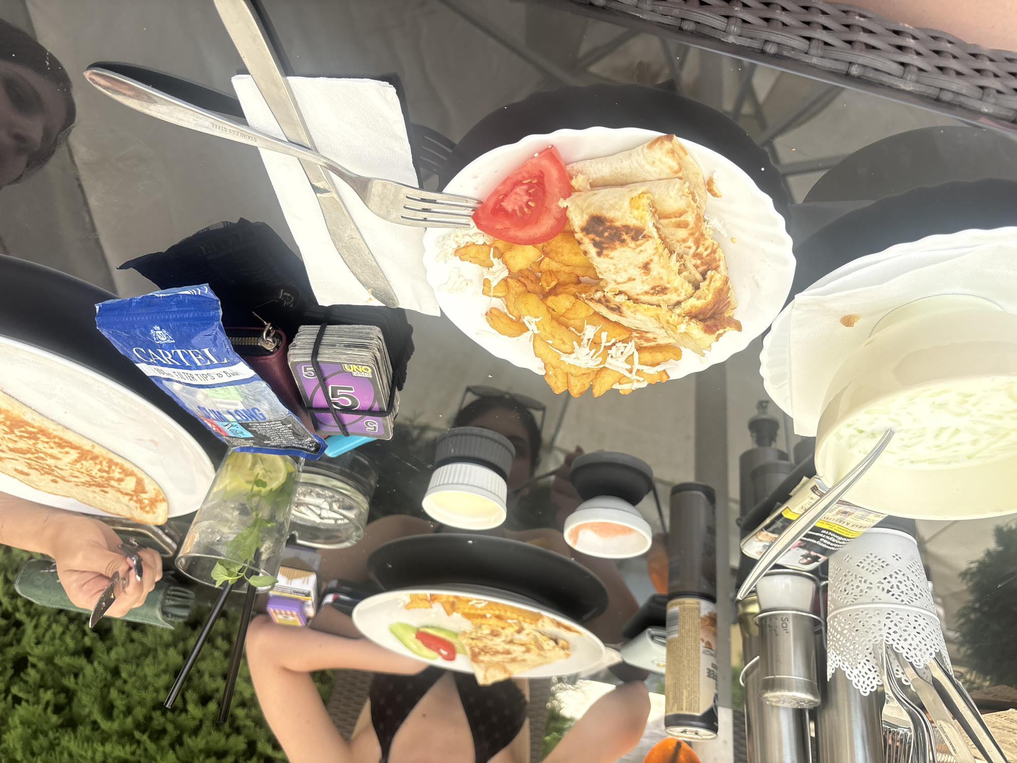 The image depicts a table setting at an outdoor dining establishment in Zverino, Bulgaria. On the table are two plates of food, one with what appears to be wraps and french fries, and another with a folded flatbread and garnishes. A white bowl filled with a green liquid, possibly soup or a cold dish, is also present. Several condiments in small white dishes are visible. In the background, two individuals are seated, one of whom is holding a drink with a straw. A can of beverage, a pack of cigarettes, an ashtray with cigarette butts, and a deck of UNO cards are scattered on the table. The table surface is black and reflective, with a wicker chair visible in the foreground. The environment suggests a casual dining setting with greenery in the background.