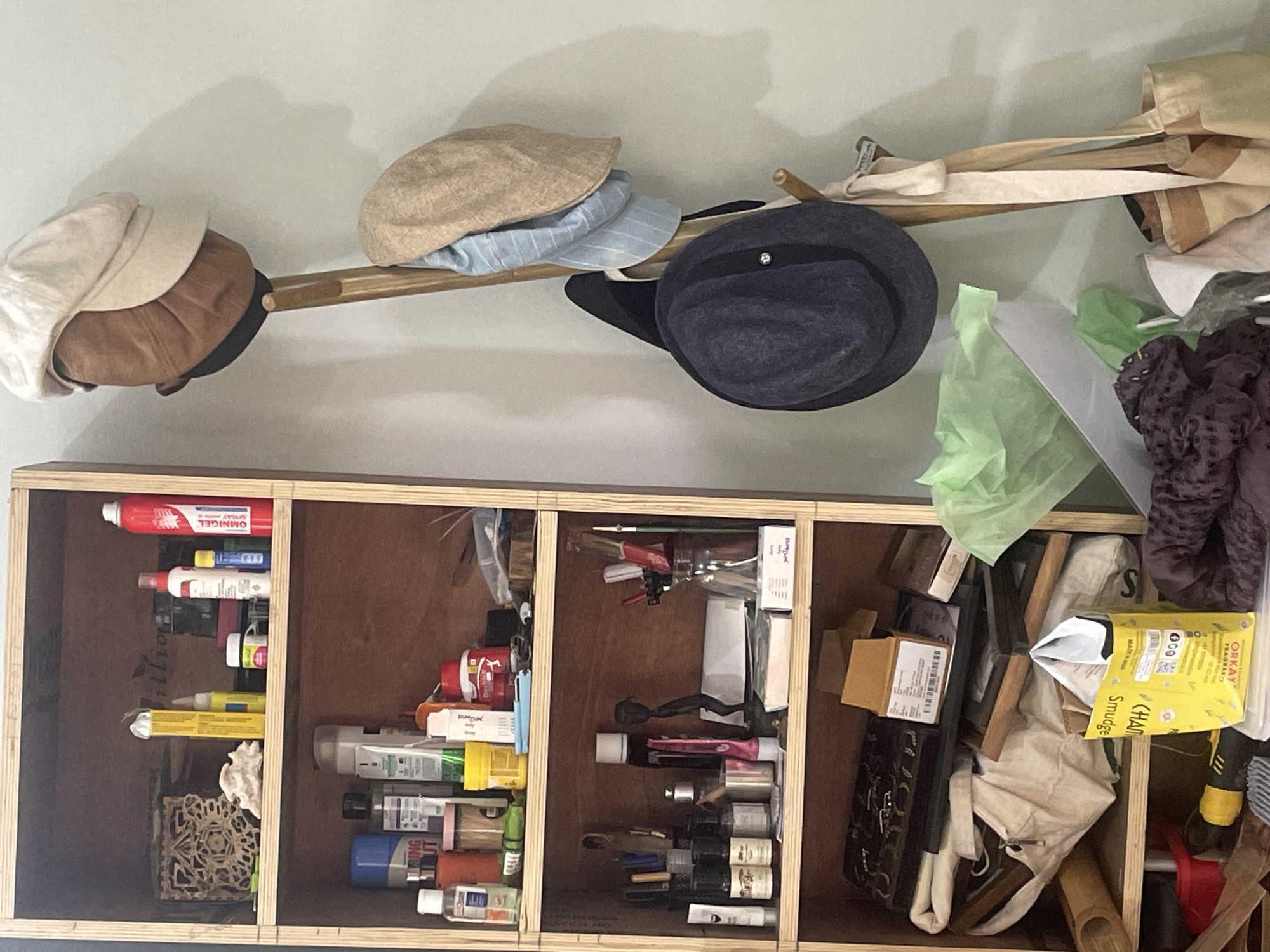The image displays a wooden shelving unit filled with various items, including bottles, containers, and boxes. On the right side of the image, a wooden hat stand holds several hats and bags. The scene appears to be indoors, possibly a room or storage area. The lighting is soft, creating shadows on the wall behind the hat stand. The overall impression is one of a cluttered but organized space. There is no indication of Bhagsu, India in the image.