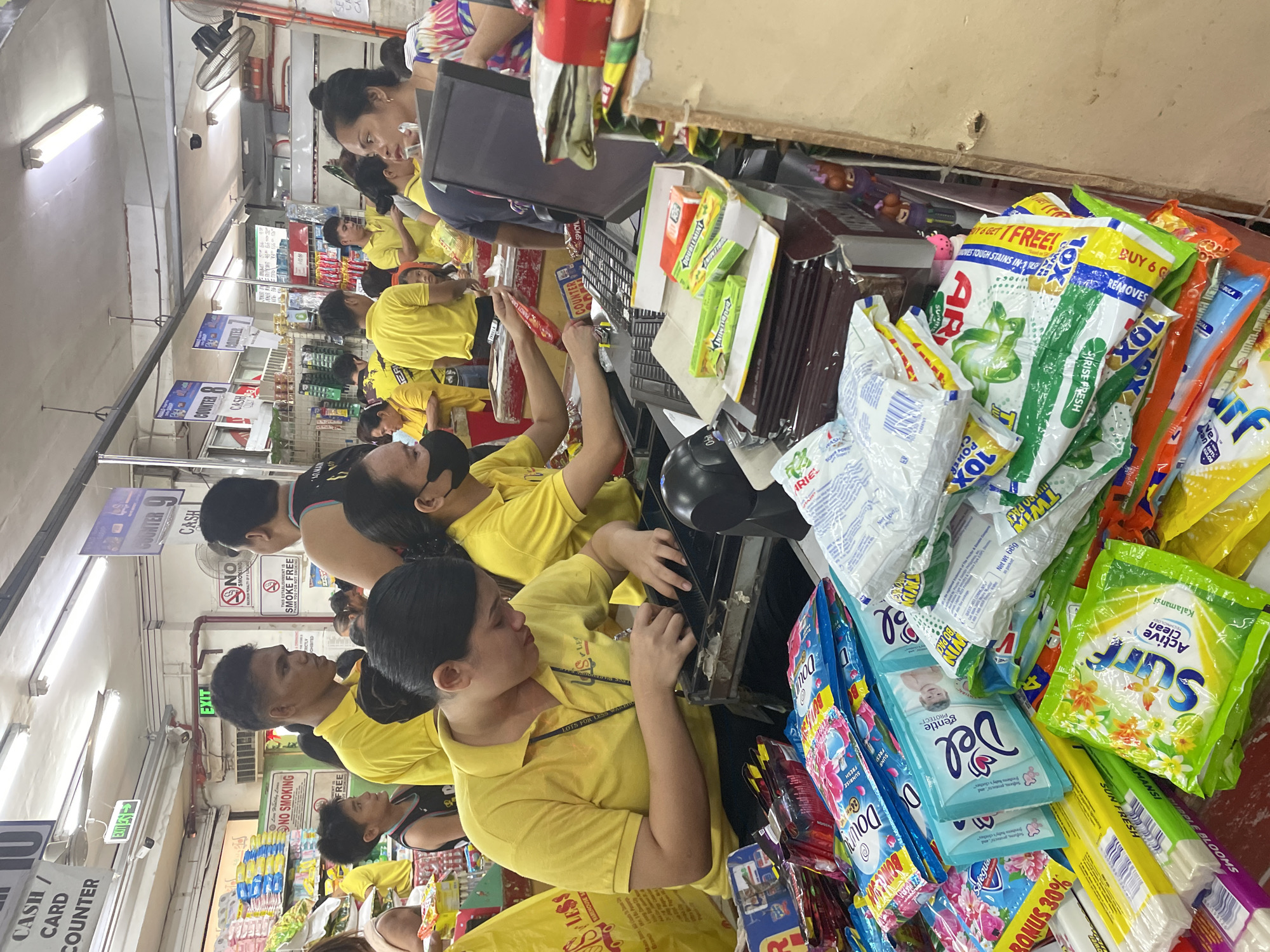 The image depicts a bustling indoor supermarket or grocery store checkout area in Davao City, Philippines. Numerous individuals are present, predominantly store employees attired in bright yellow polo shirts bearing the "Gaisano Less" logo on the left chest.
In the immediate foreground, a female cashier, identified by her yellow uniform and hair tied back, is actively processing a transaction at a cash register. Her hands are engaged with items and the cash drawer. Adjacent to her, a partially visible second female cashier is also conducting a transaction. The checkout counter in front of these cashiers is heavily stocked with various packaged consumer goods, including detergent powders (Surf Active Clean, Ariel), fabric softeners (Downy, Del), and bar soaps, indicating a large volume of purchases. A computer monitor and keyboard are positioned on the counter at the primary checkout station.
Further into the midground and background, multiple other checkout counters are operational, denoted by overhead signs indicating "COUNTER 7," "COUNTER 8," "COUNTER 9," and "CASH / CARD COUNTER 10." Several customers are visible, some queued, others interacting with staff. A male customer with a distinctive haircut, also in a yellow shirt, stands in the midground looking right. A young boy in a dark basketball jersey stands behind him. On the far right, a female customer with dark hair is partially visible near a checkout station.
The store's interior is illuminated by numerous fluorescent light fixtures attached to the ceiling. Green "EXIT" signs are mounted overhead. Additional signage includes "NO SMOKING" and "THIS ESTABLISHMENT IS SMOKE FREE." A ceiling-mounted fan is visible on the right wall. Shelves stocked with products are discernable in the background. The overall scene conveys a busy retail environment characterized by frequent customer transactions.