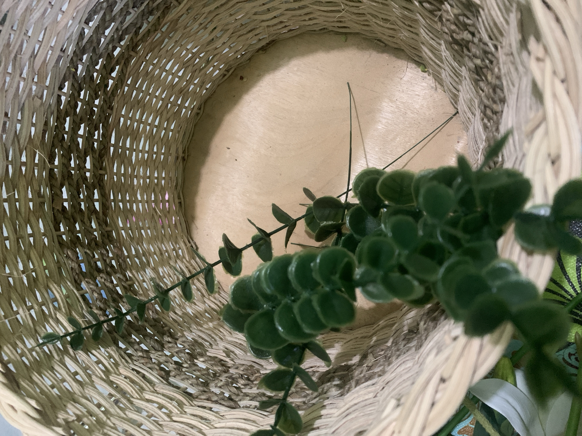 This media file shows an overhead shot looking into a woven basket. The basket is made of light-colored wicker, with a wider weave on the sides and a more tightly woven base. Inside the basket, a sprig of artificial eucalyptus leaves is placed. The leaves are a deep green, with a slightly fuzzy texture, and are arranged in clusters along thin green stems. One long, thin dark stem also extends from the plant, crossing the empty space within the basket. The interior of the basket appears to be a light-colored, smooth material, possibly wood or cardboard. The lighting suggests it is daytime, and the scene appears to be indoors, given the indoor lighting and the nature of the object. There are no people visible, nor any text. The setting is likely a home or a craft store given the presence of the artificial plant and the basket. There are no specific weather cues or strong emotional indicators. The location context of Kuala Pembuang, Indonesia, does not offer any immediately discernible visual cues within the image itself.