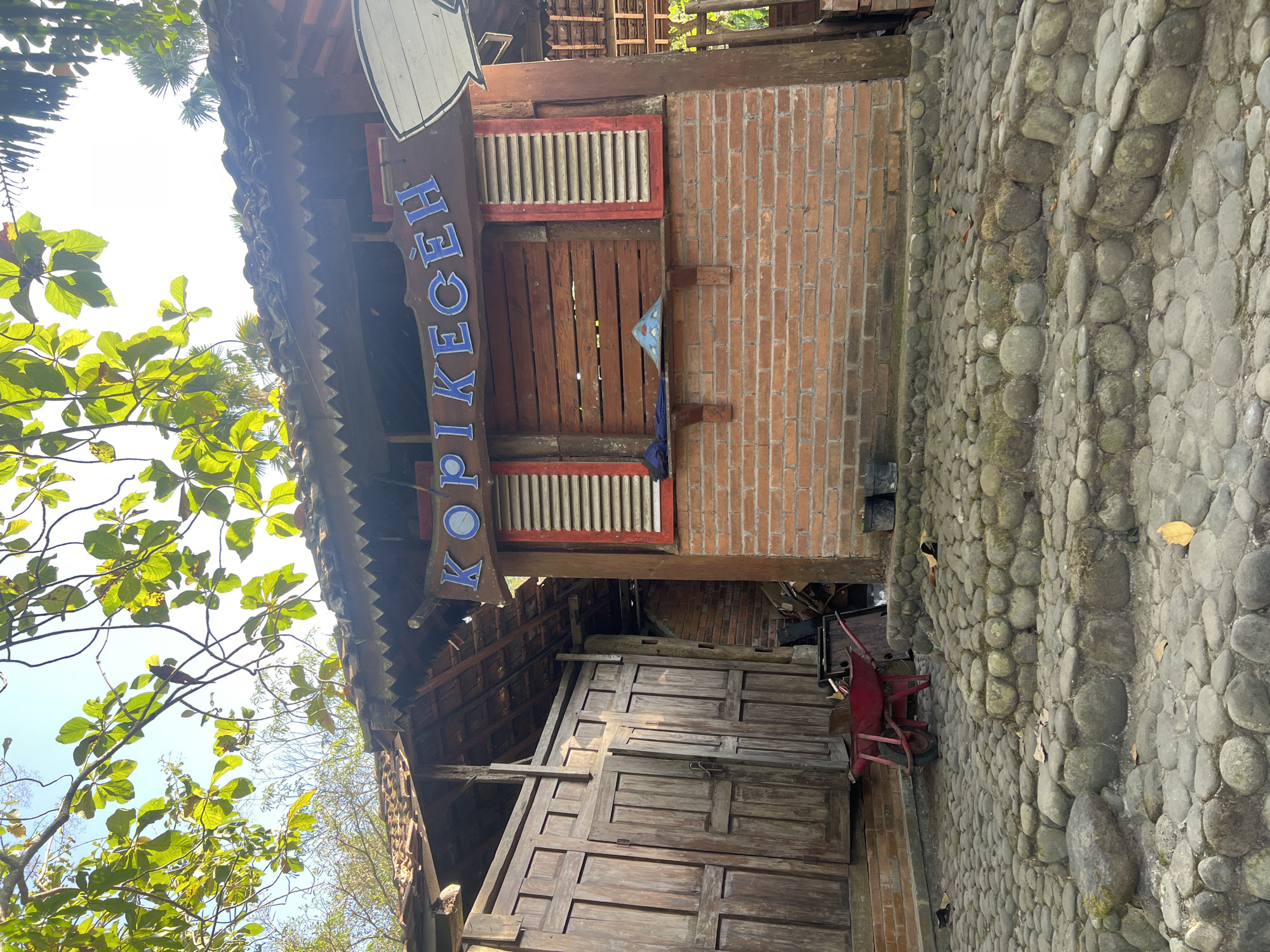 A rustic, open-air structure with a tiled roof and wooden shutters stands on a cobbled pathway in Tlogoadi, Indonesia. The building, constructed from brick and wood, features a sign with blue lettering that reads "KOPIKECEH". A red wheelbarrow is parked on the left side of the pathway, near a large wooden door. The scene is set outdoors, with trees and foliage visible in the background, suggesting a natural or garden setting. The lighting indicates it is daytime, with sunlight filtering through the leaves. There are no people visible in the image.