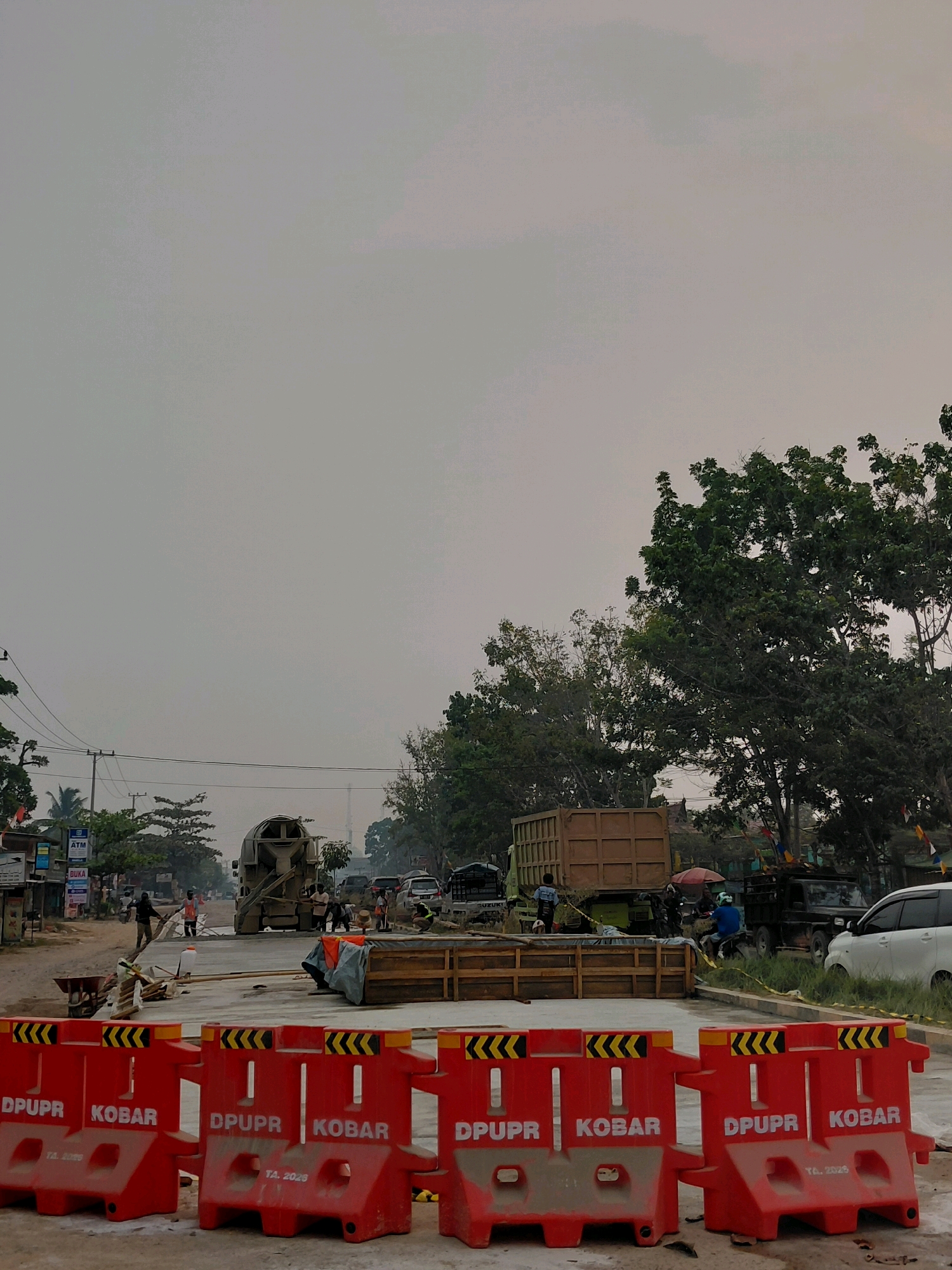 The image depicts a street construction scene in Karang Mulya, Indonesia, under an overcast sky. In the foreground, a line of bright red, plastic traffic barriers with yellow and black chevron markings is visible. Text on the barriers reads "DPUPR" and "KOBA", with "TA. 2026" printed below "KOBA".
The midground shows ongoing construction work on a road. A cement mixer truck is positioned in the center of the road, with workers around it. Further back, a large brown cargo truck with a closed trailer is parked, and other vehicles, including a black pickup truck and a white SUV, are present. Wooden formwork for concrete is also laid out on the ground. Several individuals are engaged in construction activities or observing the work.
The background features a line of buildings and shops on the left side of the road, with utility poles and power lines. Lush green trees, characteristic of the Indonesian landscape, frame the scene on the right and in the distance. Small flags are also visible attached to some of the trees and poles. The overall impression is one of active urban infrastructure development.