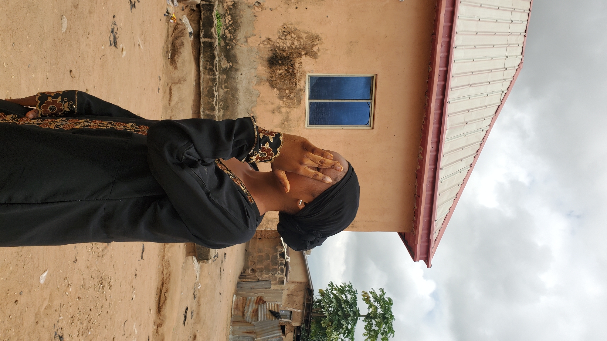 A person wearing a black hijab and a black abaya with embroidered floral patterns on the sleeves is standing in front of a tan building with a red-trimmed metal roof and a blue window. The person is covering their face with their right hand, obscuring their features. The ground is dirt, and in the background, there are other buildings, including one with a corrugated metal roof, and trees. The sky is cloudy. The scene appears to be an outdoor setting in the city of Jalingo, Nigeria, possibly a residential area.