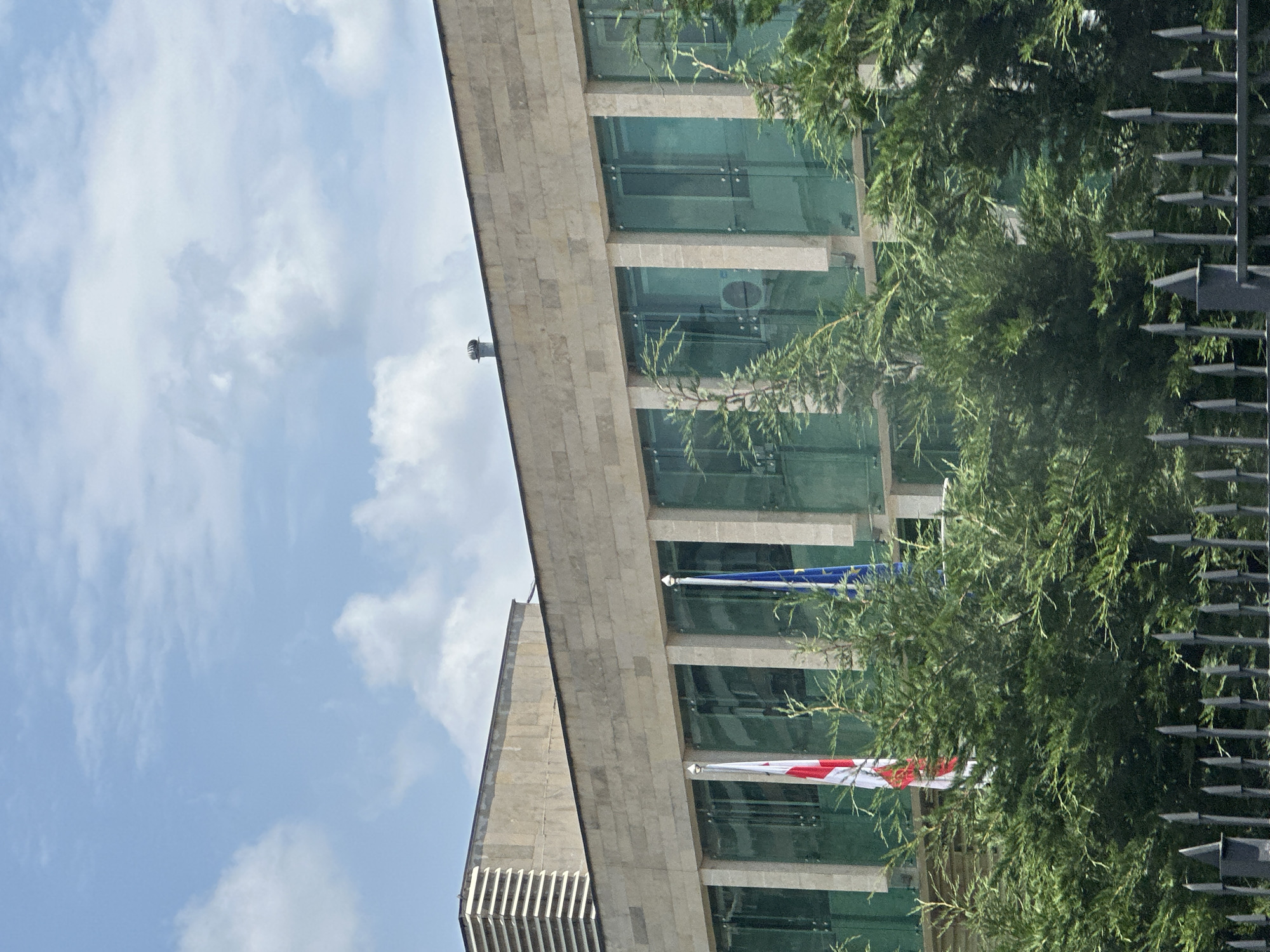 The image shows the exterior of a modern building with a facade of light-colored stone blocks and large, tinted glass windows. Two flags are visible on flagpoles in front of the building: one appears to be the flag of Georgia, and the other is the flag of the European Union. Dense green foliage, likely trees or large bushes, partially obscures the lower portion of the building and the flags. A black metal fence with pointed finials is also visible in the foreground. The sky above is blue with scattered white clouds. The overall impression is of an official or institutional building. The image does not contain any people or specific identifiable actions.