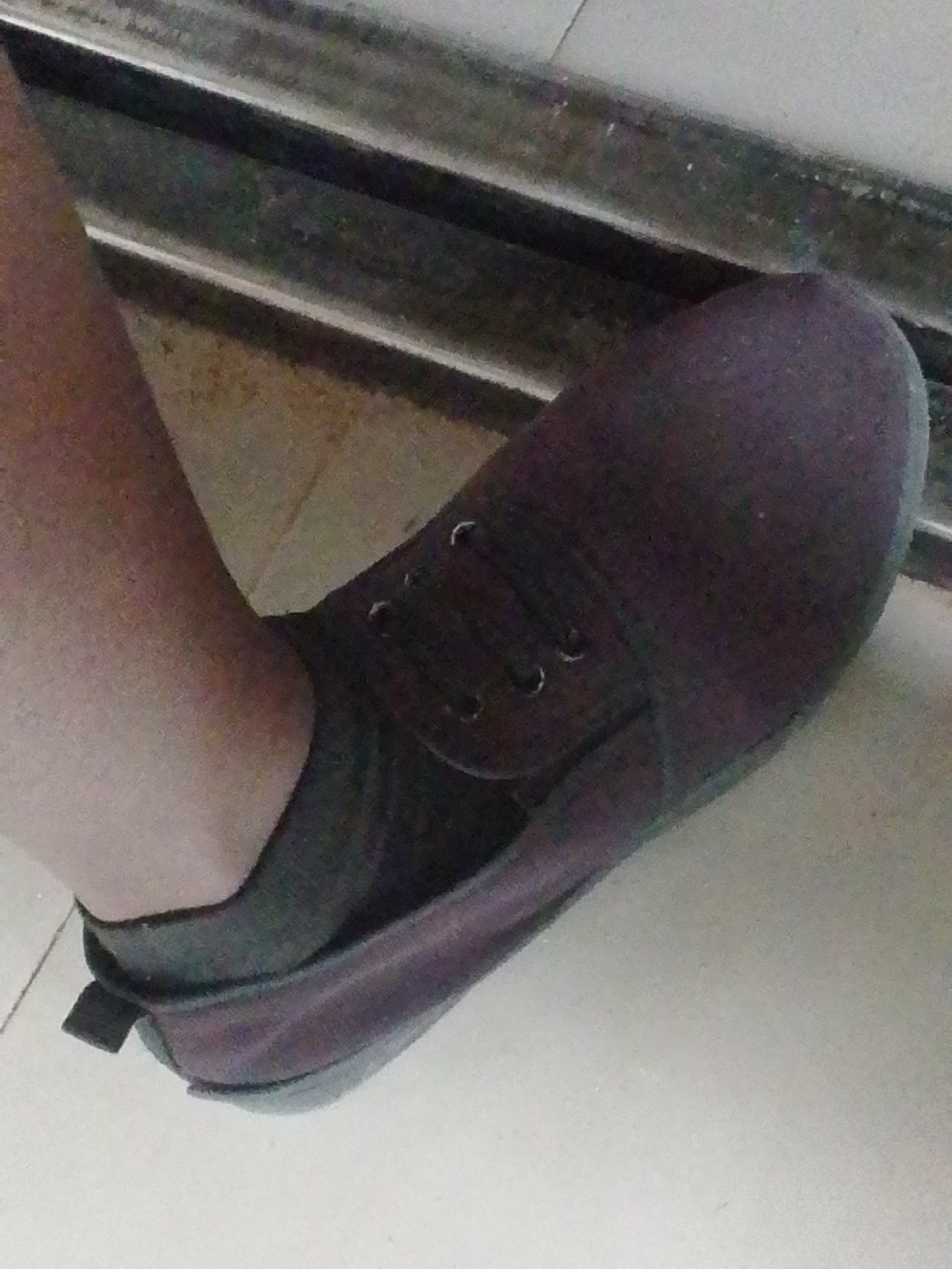 A person's foot, clad in a dark-colored shoe and a sock, is positioned on a tiled floor. The shoe appears to be a slip-on style with laces. The background shows what seems to be a doorway or a transition area, with horizontal metallic elements. The lighting suggests an indoor environment. There are no discernible landmarks to indicate the specific location within Kuson, Nigeria.