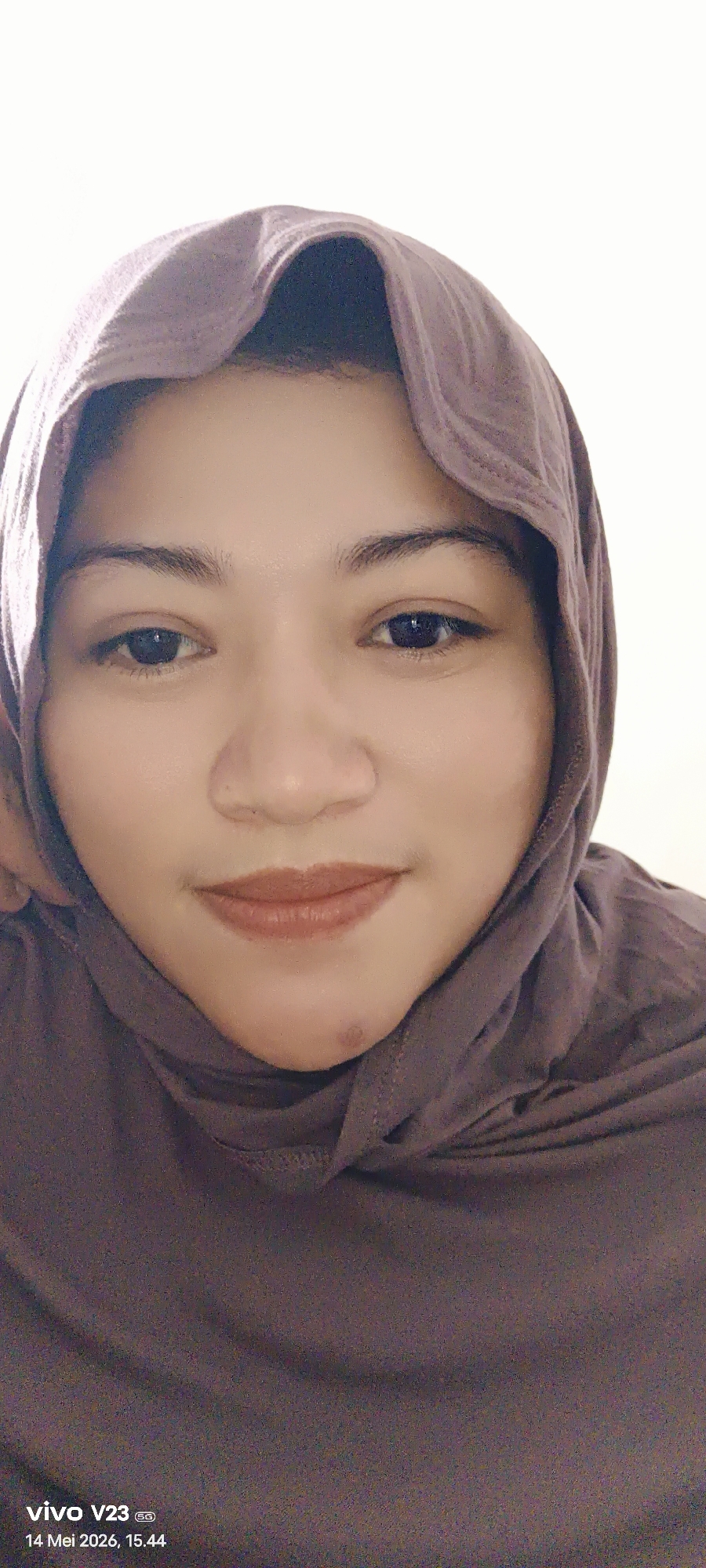 The image is a close-up selfie of a woman wearing a brown hijab. She has dark eyes, brown eyebrows, and brown lipstick. The background is a plain white wall. The photo was taken with a vivo V23 phone, as indicated by the watermark at the bottom left corner of the image. The date and time on the watermark read "14 Mei 2026, 15.44". The image does not offer sufficient details to determine the exact location in Lhokseumawe, Indonesia.