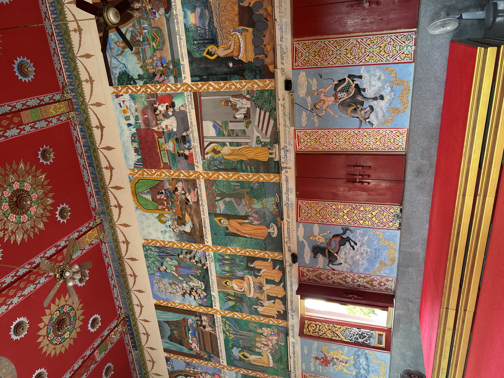 The image depicts the interior of a Buddhist temple, likely in the Ko Samui District of Thailand. The scene is dominated by a vibrant red ceiling adorned with intricate floral patterns and gilded embellishments. Two ceiling fans hang from the ornate ceiling. The walls are covered in a series of murals, each framed by decorative gold borders. These murals appear to depict scenes from Buddhist stories or teachings, featuring figures in traditional attire, some with halos, engaged in various activities such as meditation, teaching, and procession. The lower portion of the walls features large, red doors with decorative panels and further murals depicting figures on horseback and on a bull. In the foreground, a raised platform covered in red carpet with golden trim is visible, suggesting a space for religious ceremonies or seating. A white standing fan is positioned to the right of the platform. Natural light filters in from an unseen source, illuminating the space.