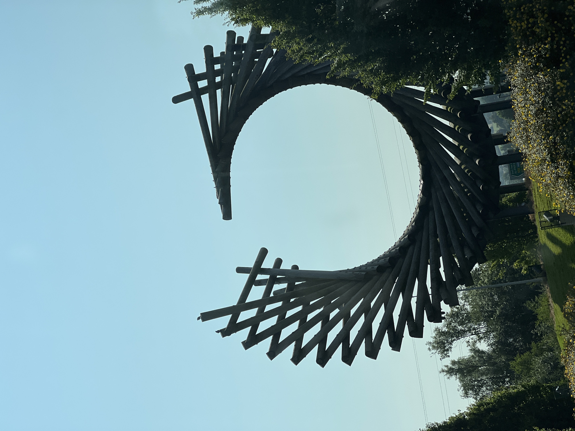 The image depicts a large, circular wooden sculpture against a clear blue sky. The sculpture is made of numerous wooden beams arranged in a spiral pattern, forming a hollow circle. Two small birds are perched on the upper left section of the sculpture. The sculpture is situated in a landscape with green trees and bushes. In the background, there is a road with a green directional sign that appears to indicate directions to "Letterkenny" and other locations. The overall setting suggests an outdoor public space, possibly a park or a roadside attraction, in or near Letterkenny, Ireland.