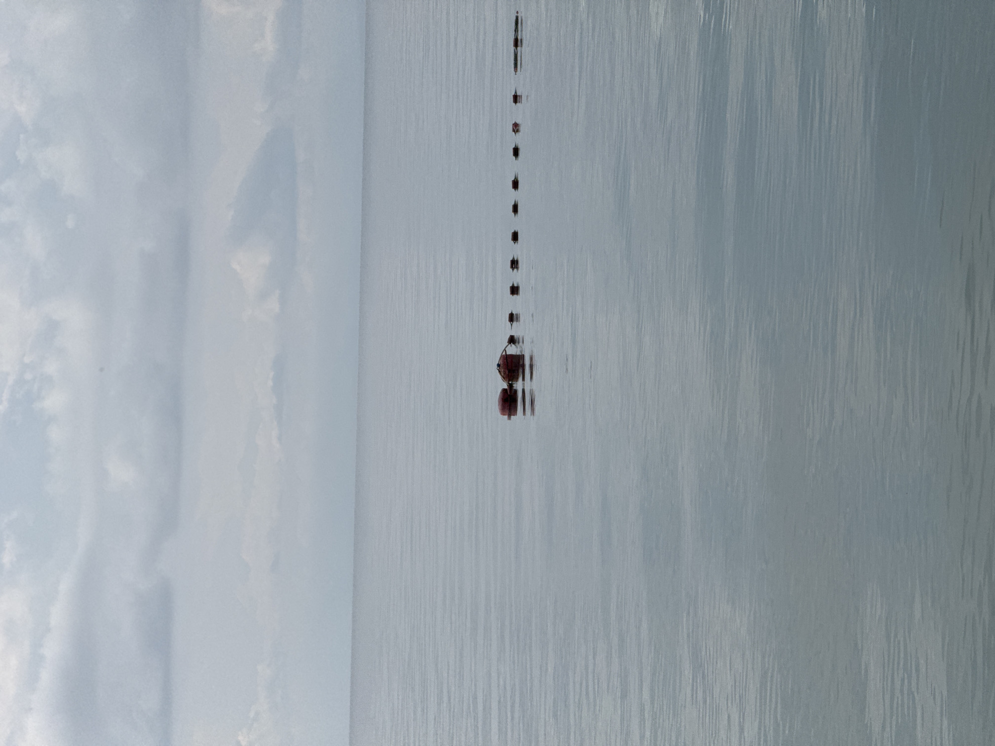 A line of red buoys stretches across a calm, gray body of water. The buoys are spaced evenly, with a larger, more rounded buoy at the left end of the line. The water is flat with subtle ripples, reflecting the overcast sky. In the distance, the horizon separates the water from a hazy, cloud-filled sky. The scene appears to be a tranquil, natural setting.