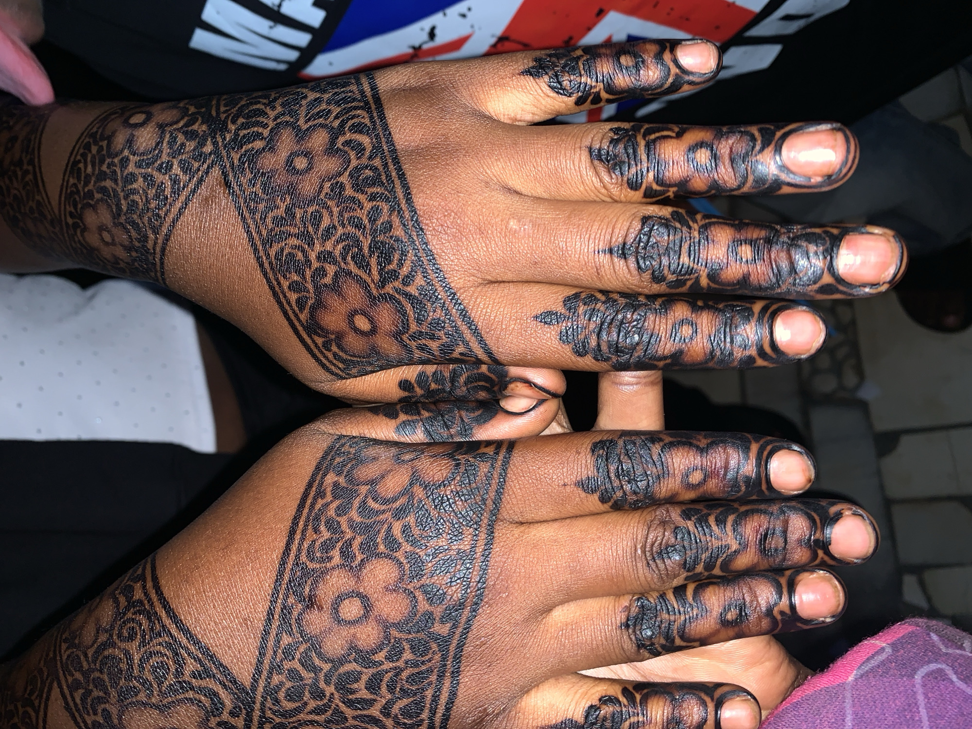 The image shows a close-up of two hands adorned with black henna tattoos. The henna designs cover the backs of the hands and extend up the wrists, featuring intricate floral and geometric patterns. The fingers are also heavily decorated with dark henna. The skin tone of the hands is dark brown. The background is blurred, but suggests an indoor setting with some clothing visible, including what appears to be a white collared shirt and a dark t-shirt. The overall impression is of freshly applied henna. The location is identified as New Stadium, Nigeria.