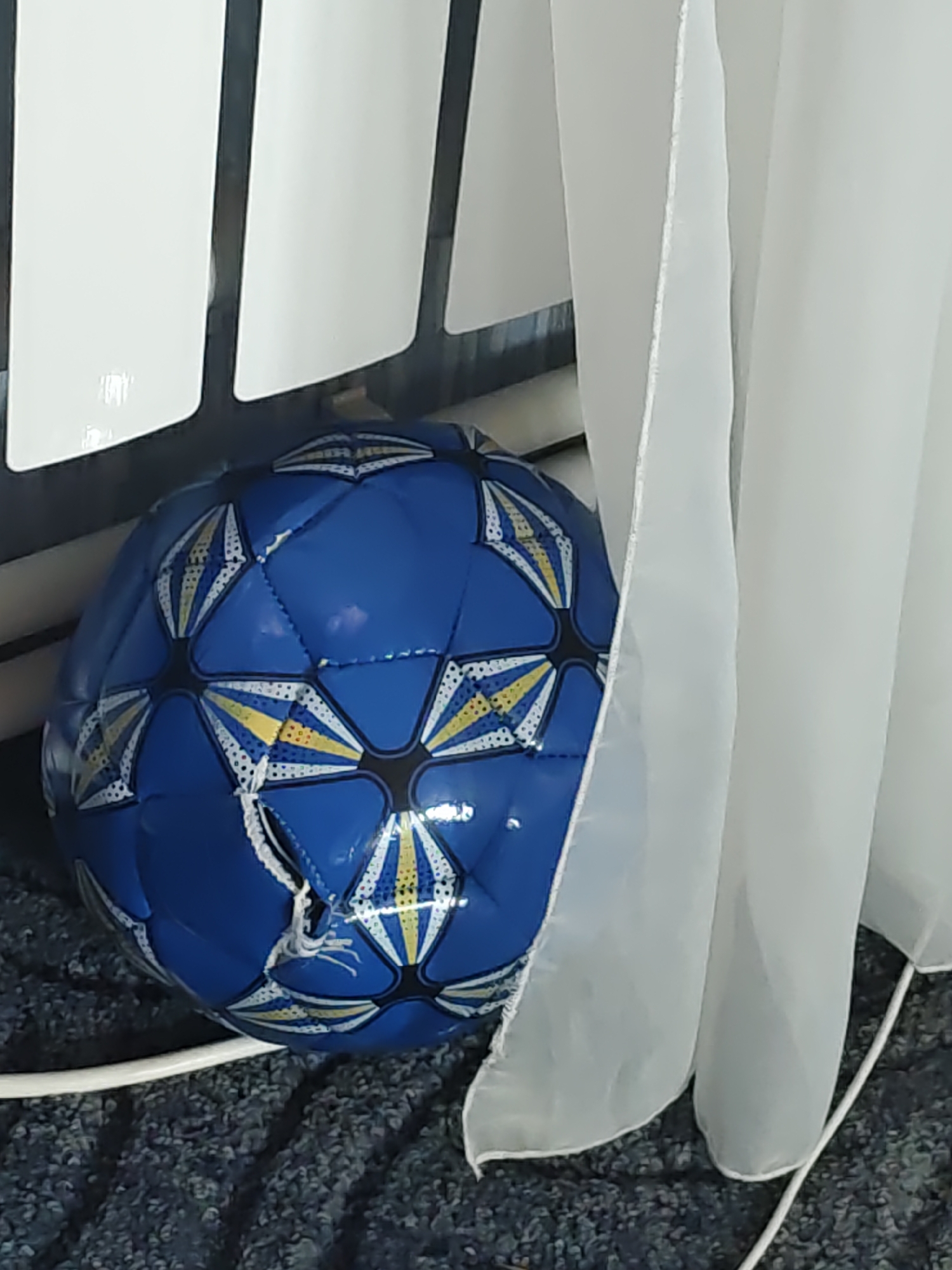 A blue soccer ball with white and yellow hexagonal patterns is resting on a dark, textured surface. The ball is partially obscured by a sheer white curtain, which hangs to the right of the frame. In the background, a dark surface with vertical white rectangular elements is visible. The scene is likely indoors.