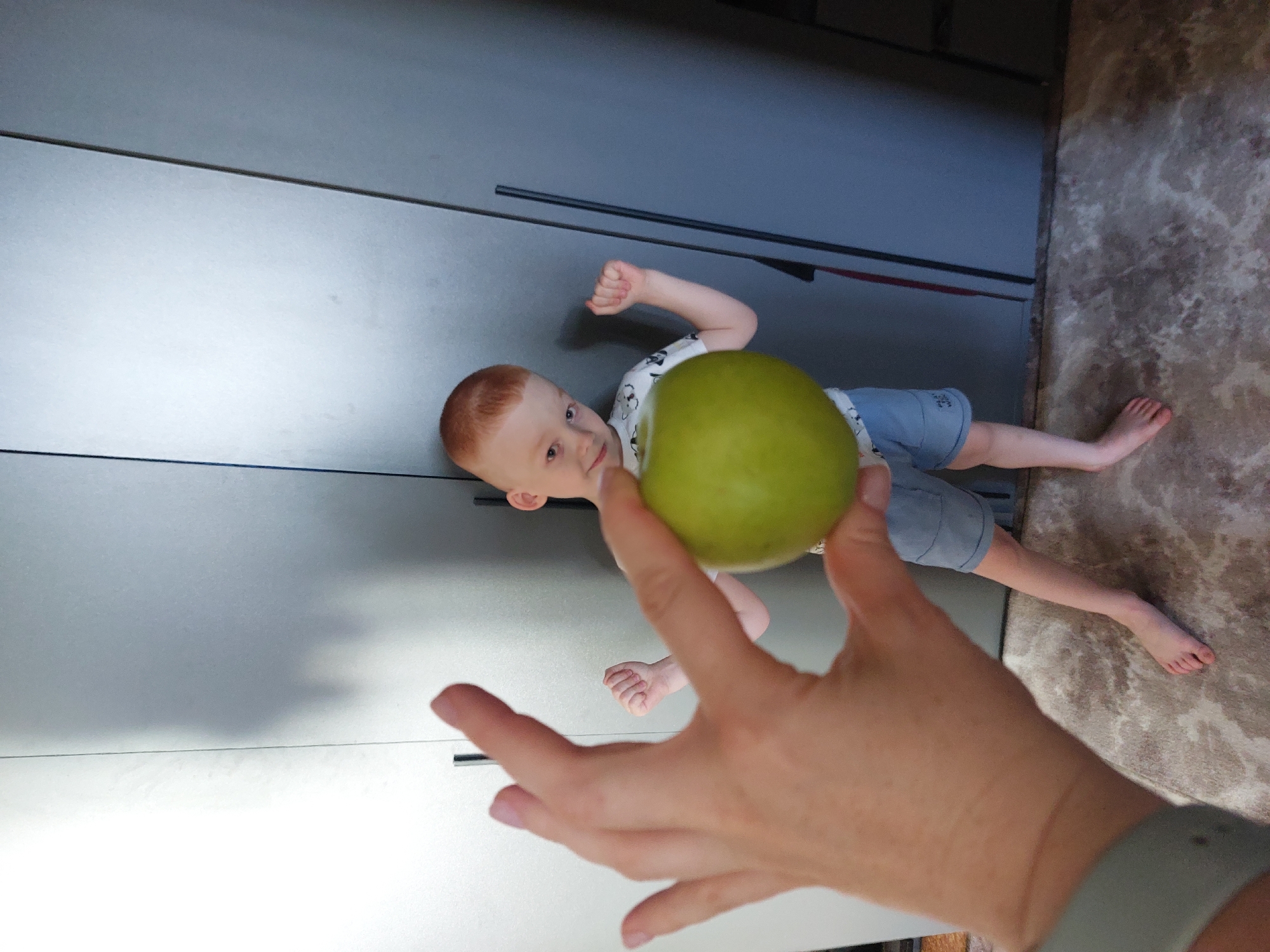 A young boy with red hair, wearing a white t-shirt with a cartoon print and blue shorts, is lying on the floor. He is looking at the camera and flexing his bicep. In the foreground, a person's hand is holding a green apple, obscuring part of the boy. The background consists of a large, gray, modern wardrobe with vertical handles. To the right of the wardrobe, a textured, beige and gray floor is visible. The scene appears to be indoors, likely in a home. The lighting suggests it could be daytime. No text is visible in the image.