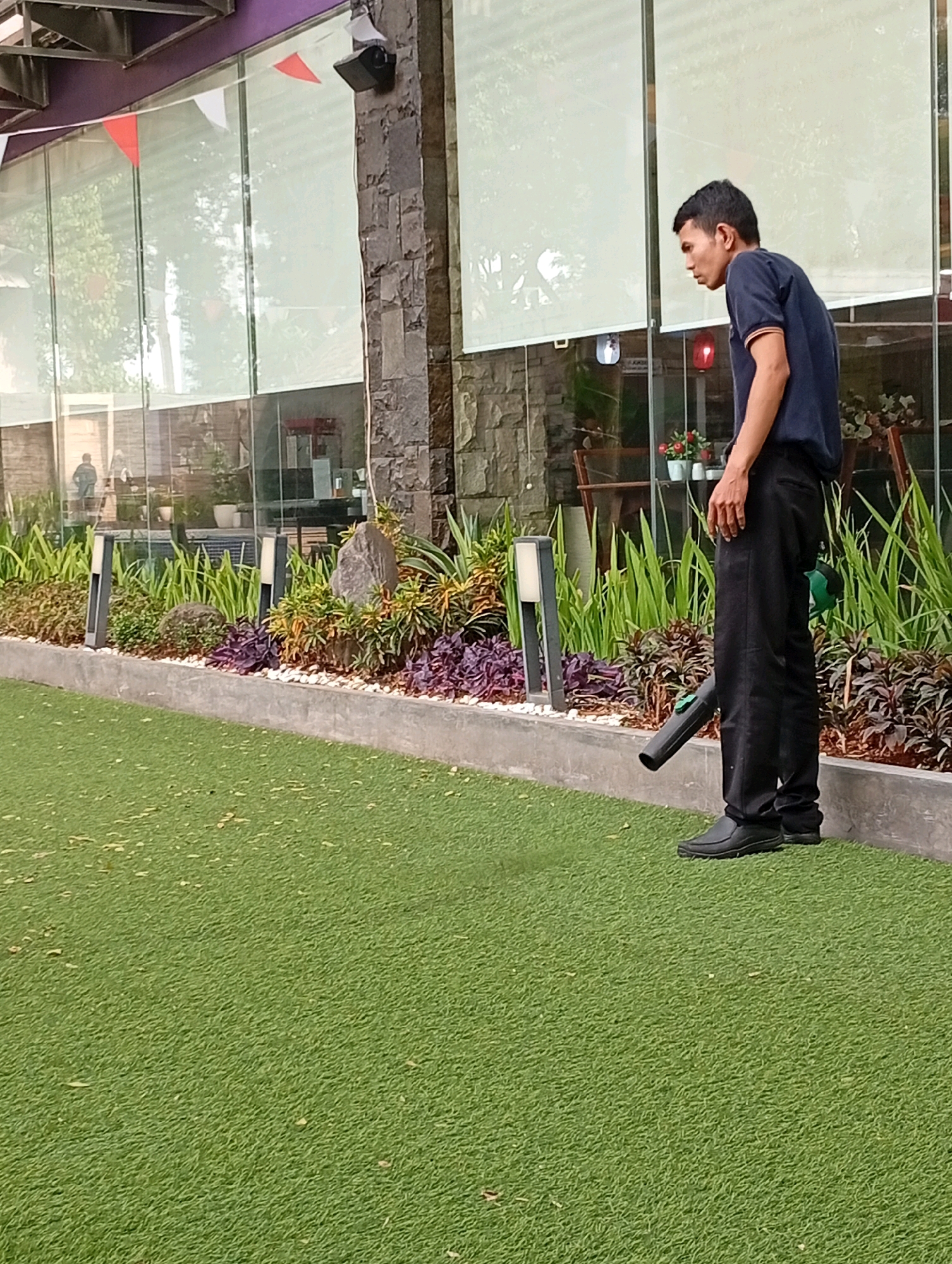 A man wearing a dark blue polo shirt and black pants is standing on a green artificial turf area, holding a green leaf blower at a downward angle. He is facing towards the right of the image and looking down. Behind him is a planter box filled with various green and purple plants, with decorative rocks and modern-style lamps. Beyond the planter box is a building with large glass windows, a stone facade, and a purple overhang. The scene appears to be an outdoor area, possibly a cafe or restaurant, in Binong, Indonesia.