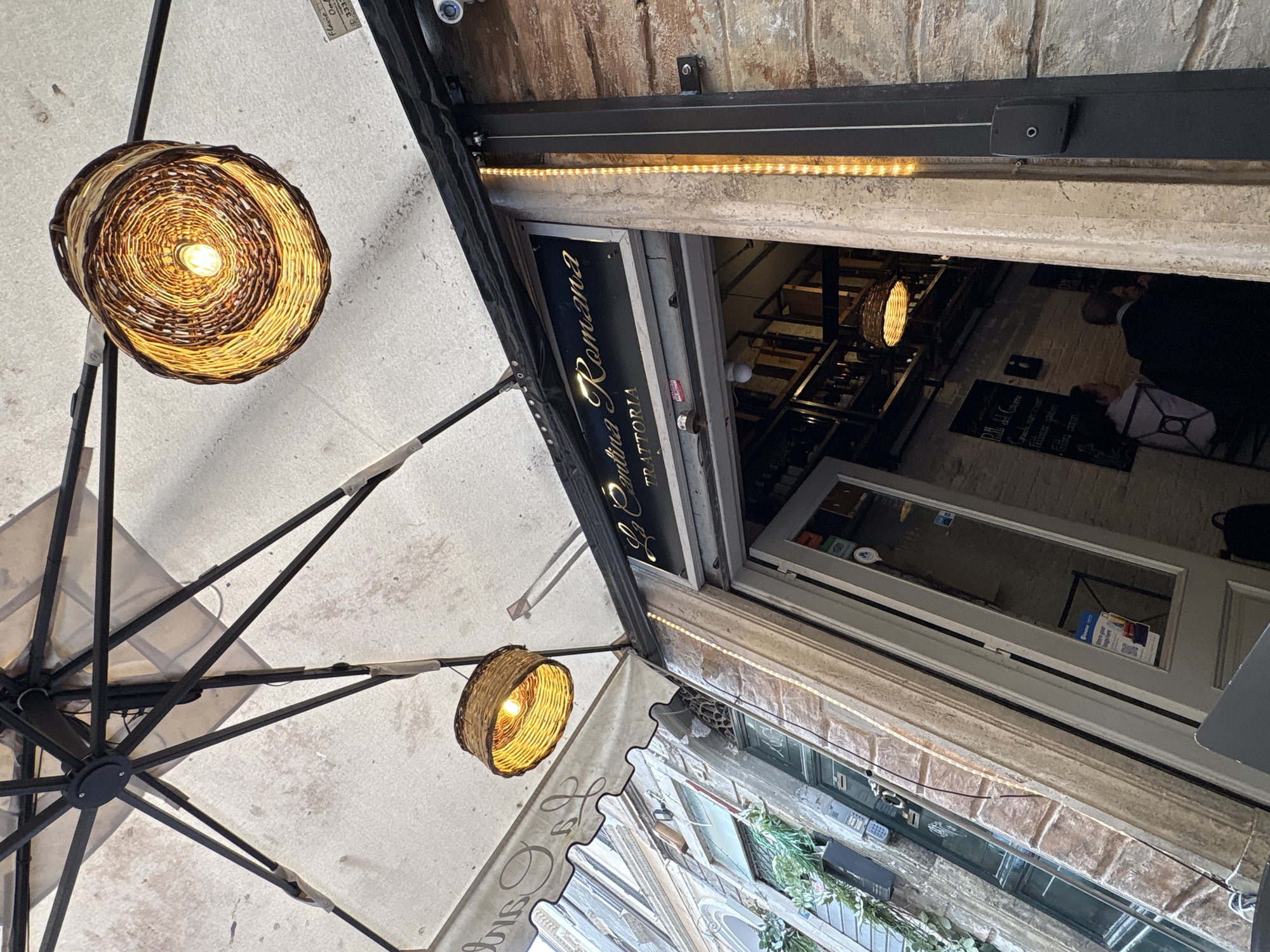 This vertical shot, taken from a low angle looking upwards, captures the vibrant entrance of a trattoria, likely in Rome given the visible Italian text. In the foreground, a large, light-colored outdoor umbrella with a dark metal frame dominates the left side of the frame, providing shade. Two decorative lamps with woven, basket-like shades and warm glowing bulbs are suspended, one prominently in the upper left, hanging from the umbrella's structure, and another partially visible below it.
The main subject is the entrance to "La Bottega Romana" TRATTORIA, clearly identified by a black sign with gold lettering above the doorway. An illuminated LED strip runs along the top of the entrance frame, casting a bright light. The building's facade features textured stone or brickwork above the entrance, and a security camera is visible on the wall.
Through the glass door of the trattoria, the dimly lit interior shows hints of another similar lamp and the faint presence of individuals, suggesting patrons. A small sign with a QR code is visible inside the door. On the right side, below the main trattoria entrance, another old building's facade with windows and some greenery adds to the urban street scene. The combination of artificial lighting and ambient light suggests either a shaded area or late afternoon/early evening. The overall atmosphere is inviting and characteristic of a European city.