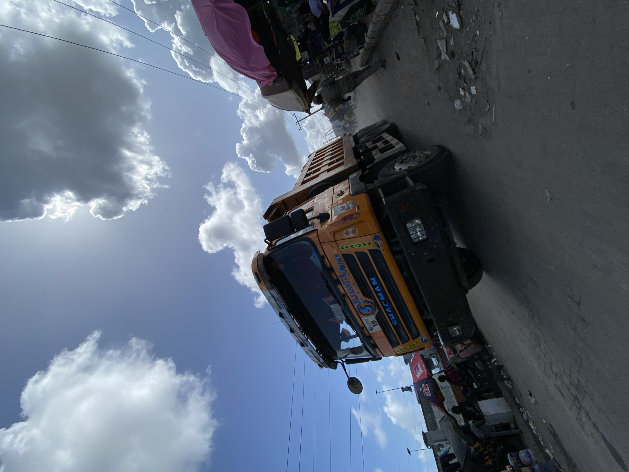The image depicts a street scene in Lagos, Nigeria. A yellow dump truck with "Alexander 4800" and "Shacman" markings is prominently featured on the right side of the frame. The truck is angled towards the viewer, suggesting it is moving along a paved road. The sky above is partly cloudy with a mix of blue sky and large, white clouds. Along the roadside, there is evidence of commerce with stalls and miscellaneous objects clustered together under a pink umbrella. There is evidence of litter and debris on the road and along the roadside. Buildings and overhead power lines are also visible. People can be seen interacting in the market area.