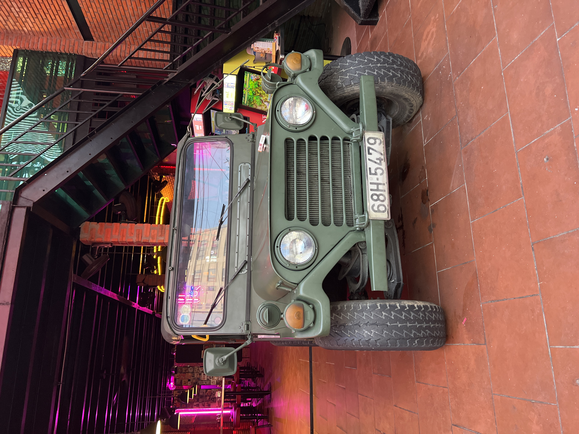 The image depicts an olive green, military-style utility vehicle, possibly an older model jeep, serving as a prominent display piece indoors in Bac Nha Trang Ward, Vietnam. The vehicle is positioned on a reddish-brown tiled floor, facing the viewer head-on, showcasing its large circular headlights, a vertically slatted grille, and robust, treaded tires.
The setting suggests a commercial establishment, likely a bar or restaurant, characterized by its industrial-chic decor. To the left and above the jeep, a black metal staircase ascends alongside an exposed brick wall. The background behind the vehicle is dimly lit, predominantly by vibrant neon lights in shades of pink, purple, and yellow, creating an atmospheric and modern ambiance. Several electronic screens are also visible in the background, adding to the venue's contemporary feel.
The strong artificial lighting, particularly the neon glow, indicates that the photograph was taken in the evening or night, or within an interior space with controlled lighting. No people or specific activities are visible, emphasizing the static display of the vehicle within its decorated environment.
A white license plate is attached to the front of the jeep, clearly displaying the text: "68H-5479".