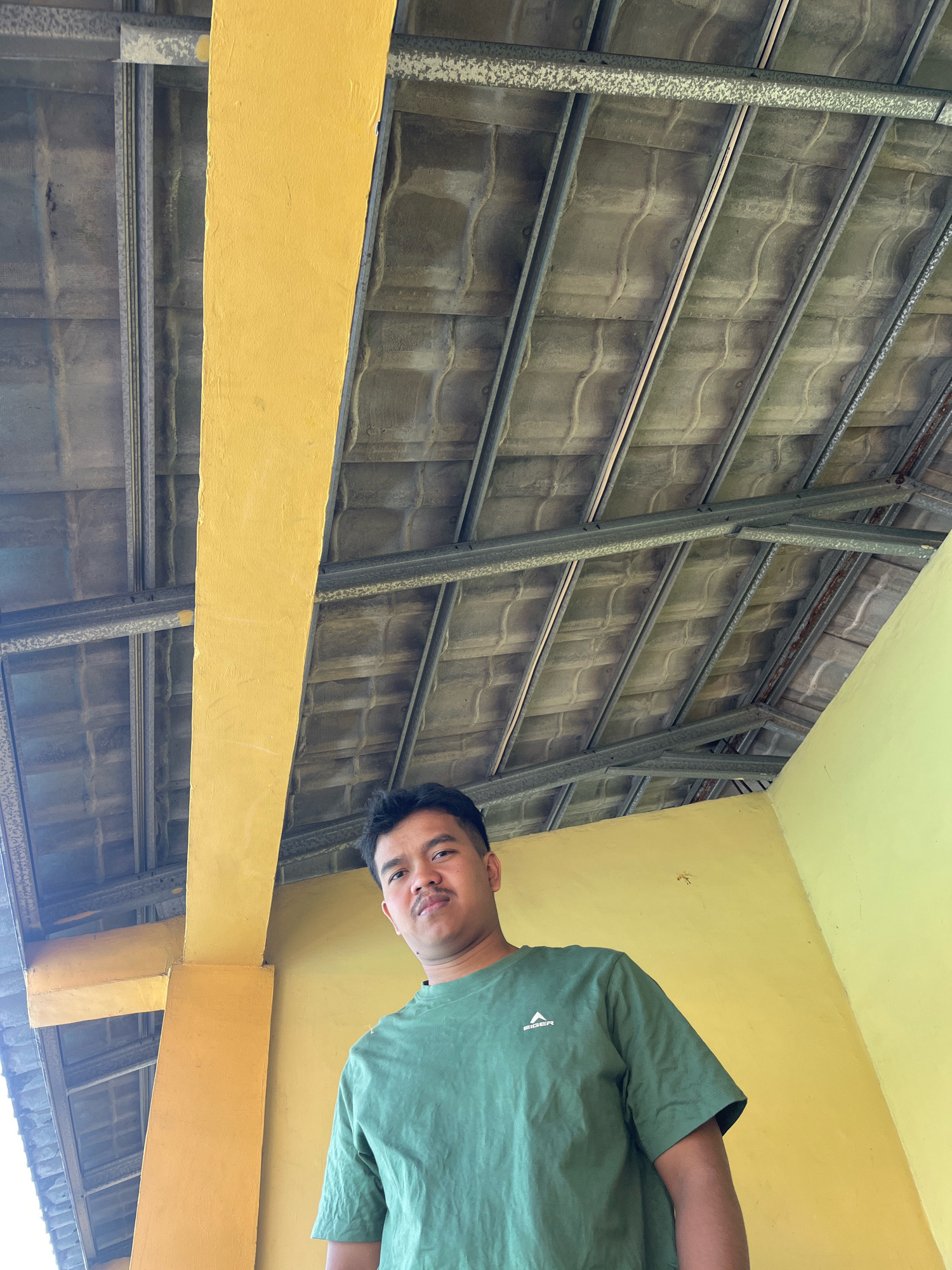 The image shows a man standing outdoors, looking up at the camera. He is wearing a green t-shirt with a white logo on the left side of the chest. Behind him, a yellow pillar extends upwards, and a section of a roof with grey tiles and metal framing is visible. The man has short, dark hair and a mustache. The overall scene suggests an open-air structure, possibly a building or a covered walkway, in a tropical or subtropical climate, given the materials and the man's attire. The lighting indicates it is daytime, likely with the sun positioned overhead or slightly to the side.