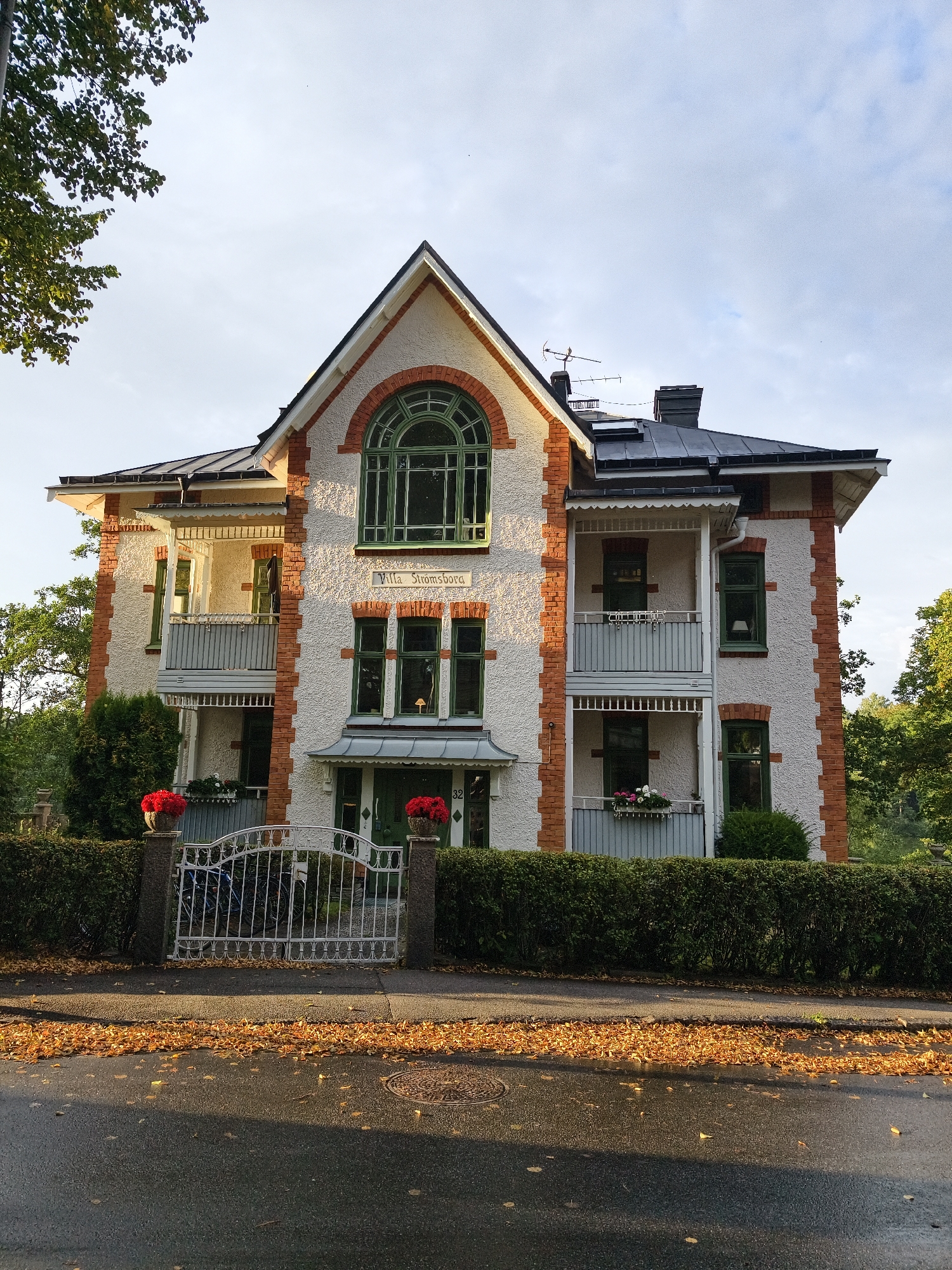 The image shows "Villa Strömsborg" located at Skogsvägen 32, Norrköping, Sweden. It is a two-story house with a white textured facade, red brick accents, and a dark pitched roof. The house features several green-framed windows and two balconies. The property is surrounded by a dense green hedge and accessed by a white ornate metal gate. Fallen orange leaves are scattered on the asphalt road in front of the hedge and gate. The sky above is partly cloudy, and sunlight casts shadows on the road. There are no people or animals visible in the image.