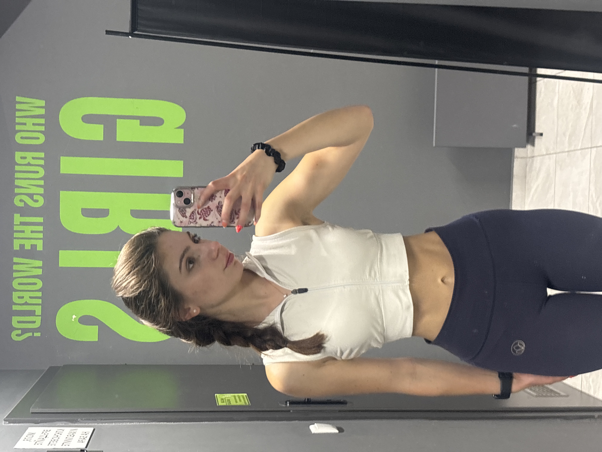 A young woman stands in front of a gym mirror, taking a selfie. She is wearing a white zip-up workout top and dark blue leggings. Her hair is braided and she is wearing a black scrunchie on her wrist. She is holding a cell phone with a turtle-patterned case. The reflection in the mirror shows the gym's motivational graffiti and interior. The exact location in Varna, Bulgaria, cannot be determined from the image.