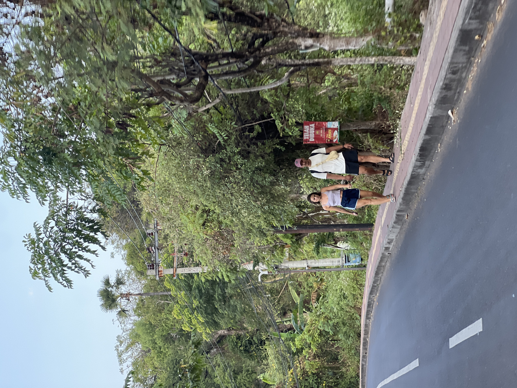An outdoor, medium shot captures a couple walking along a paved sidewalk next to a road, with dense green foliage and trees lining the background. The couple, walking from left to right, appears to be in Abang, Indonesia, suggested by a red and white sign in Indonesian near them that reads "TELAH DIBUKA" (meaning "Opened") and "AMAN" (meaning "Safe"). The man on the right wears a white t-shirt, black shorts, a cap, and a cross-body bag. The woman on the left wears a light purple tank top and blue shorts. They are walking on a sidewalk with a reddish-brown brick pattern. The road in front of the sidewalk has white lane markings. Electrical poles and wires are visible amidst the trees in the background, which also include palm trees. The sky is a light blue with a hazy quality.