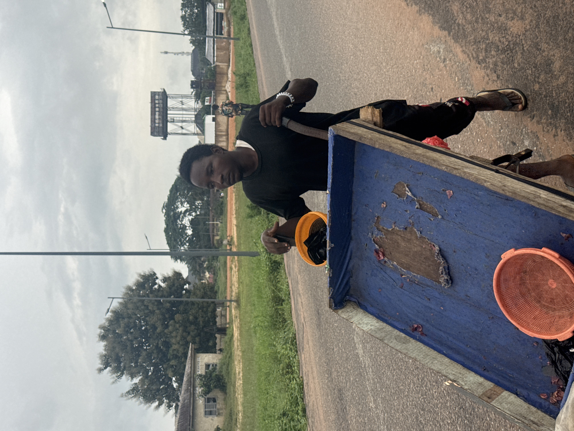 A young African man is pictured pushing a cart laden with what appears to be goods, possibly fish, along a road in Ihiagwa, Nigeria. The man wears a black t-shirt, black pants, and flip-flops. He is holding a bent metal rod connected to the cart, using it to propel the cart forward. An orange plastic bucket, partially filled with what looks like plastic bags, is visible in his right hand. The cart's surface is covered with a tattered blue tarp, and scattered on it are pieces of red material, possibly remnants of the goods. An orange plastic basket sits on the tarp. In the background, lush green vegetation lines the side of the road, with a few buildings and trees visible. A large water tower stands in the distance. The sky is overcast, suggesting a cloudy day. Another person, indistinctly visible, is walking in the background. The road appears to be paved with asphalt.