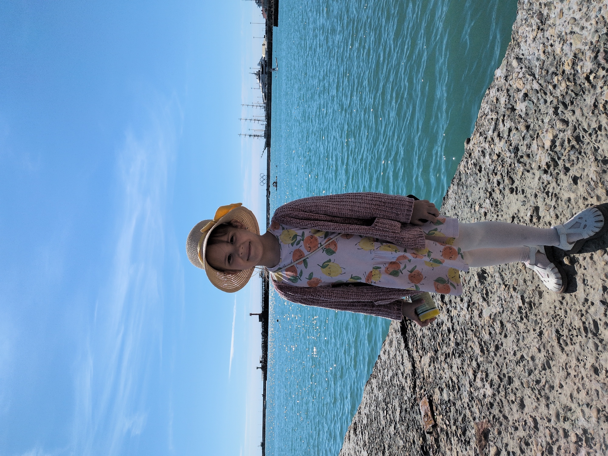 A young girl is standing on a textured concrete pier next to the sea, under a clear blue sky. The image is originally oriented vertically, with the girl positioned on the right side, looking towards the camera.
The girl wears a straw hat, a light-colored dress patterned with lemons and oranges, a pink knitted cardigan, white tights, and white sandals. She holds a small yellow-green object in her left hand and displays a subtle smile.
To her left, the turquoise sea glistens with sunlight reflecting off its rippling surface. In the background, a distant shoreline is visible, featuring various structures that suggest a port or urban area, including several ships and boats, one of which is a large sailing ship with multiple masts. A faint lighthouse or beacon can also be seen further out. The sky above is a bright blue with scattered, wispy white clouds. The rough, pebbly texture of the concrete pier occupies the foreground and extends to the girl's right. The scene conveys a sunny and pleasant day by the water in Sochi, Russia. There is no visible text in the image.