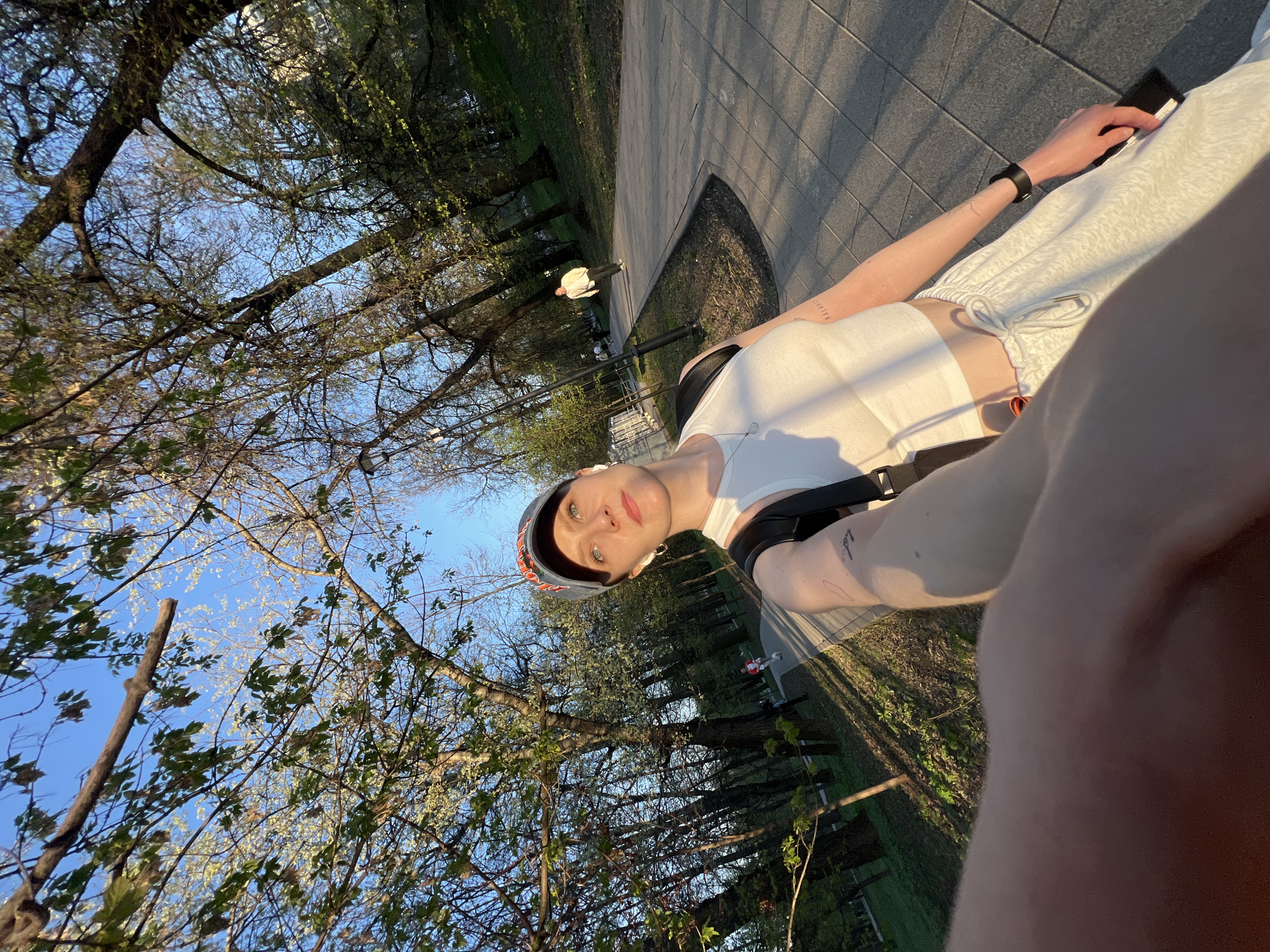 A low-angle, rotated, outdoor shot captures a person wearing a helmet and white athletic clothing seated on a wheelchair along a paved pathway. The person is looking towards the camera. Their right hand is visible holding a black object, possibly a phone. The person appears to be wearing a watch on their left wrist. A black harness is secured around the person's torso. Trees with budding leaves are visible to the side of the pathway, along with a park setting. In the background, another individual wearing light clothing is visible on the pathway. The location is assumed to be Unknown, Unknown.
