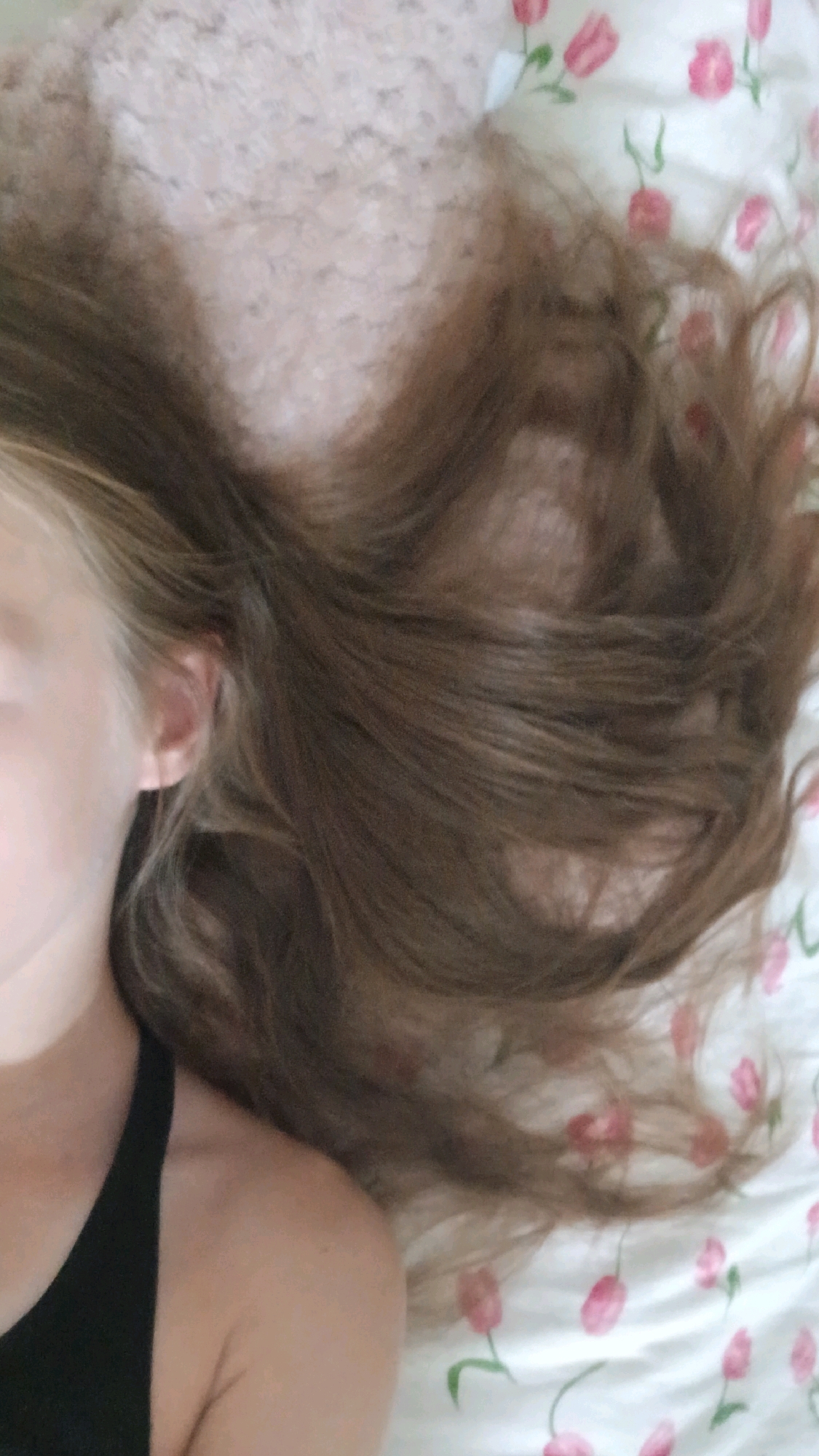 The image displays a close-up, overhead view of a person's head and shoulder, lying down. The person has long, light brown hair spread across a floral-patterned fabric, possibly a bedsheet or blanket, which features pink tulips. A portion of the person's face, including an ear, is visible on the left side of the frame. The person is wearing a black garment, likely a top or dress, visible on the left shoulder. The overall lighting suggests an indoor setting. There is no identifiable information within the image that would indicate its location in the city of Haivoron, Ukraine.