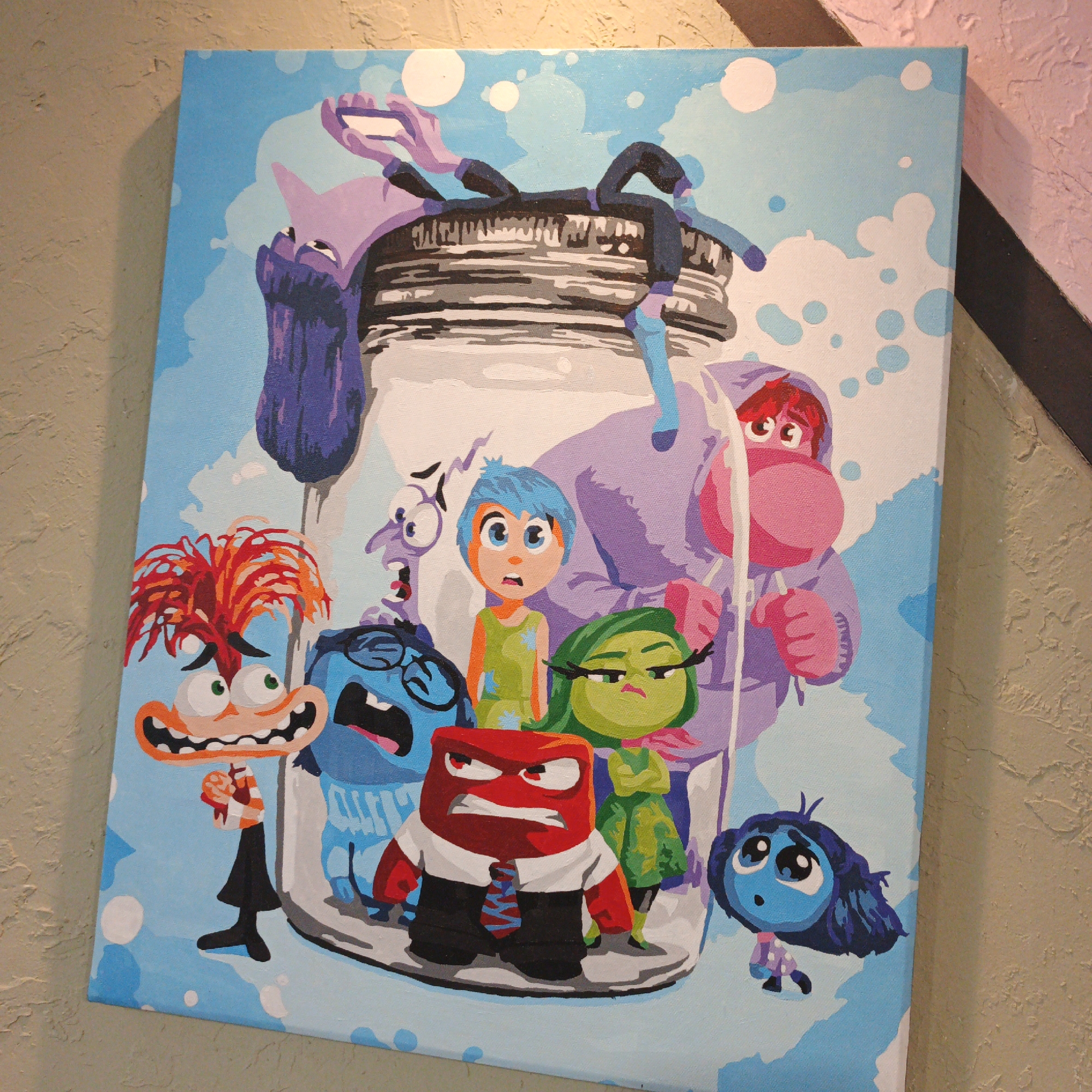 The image is a painting on canvas featuring characters from the Pixar movie "Inside Out." The characters are depicted inside a large glass jar, with a light blue, cloudy sky as the background. The characters include Joy (blue hair), Sadness (blue skin), Anger (red), Fear (purple), Disgust (green), and Bing Bong (pink elephant-like creature). Joy is in the center, looking distressed. Fear is to her left, with a terrified expression. Sadness is to Joy's right, looking concerned. Anger is in the foreground, wearing a suit and tie, with a furious expression. Disgust is to Anger's right, looking unimpressed. Bing Bong is a large figure in the background, partially obscured by the jar. The painting is displayed indoors, leaning against a textured, light brown wall. The lower right corner of the image shows a sliver of a pink wall and a dark, diagonal line, possibly part of the room's decor or another object. The painting is lit from above, casting a slight shadow. The location in Vyshgorodok, Russia is not visible in the image and cannot be determined.