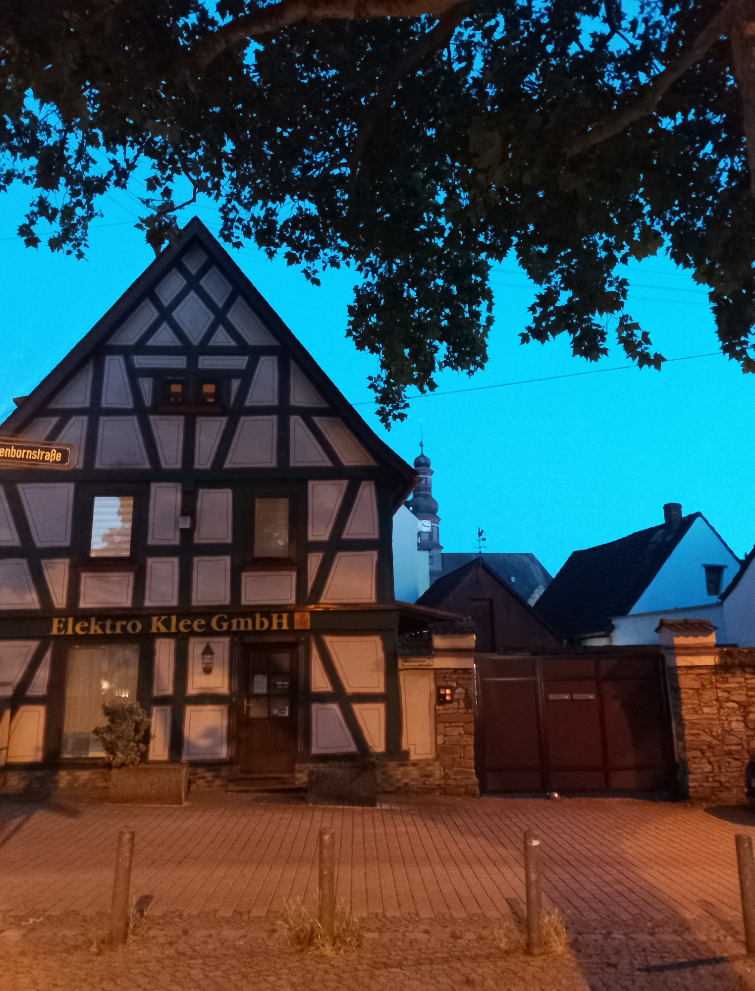 An exterior, medium shot captures a street-facing view of a historic building in a town at dusk. The building, with its distinctive half-timbered facade, stands under a deep blue sky. A large tree with dark foliage partially obscures the upper portion of the frame and the roofline of the building. The facade features dark wooden beams forming geometric patterns against a lighter background, with several windows visible. A sign with German text, "Elektro Klee GmbH," is prominently displayed on the lower part of the building. To the left, another sign reads "ßenbornstraße". Adjacent to the building, a large, dark metal gate is set into a stone wall. In the background, the spire of a church tower with a clock face is visible, along with the rooftops of other buildings. The ground in front of the building is paved with brickwork and lined with short, weathered posts. The lighting suggests the scene is illuminated by ambient light and possibly some interior lighting from the building.