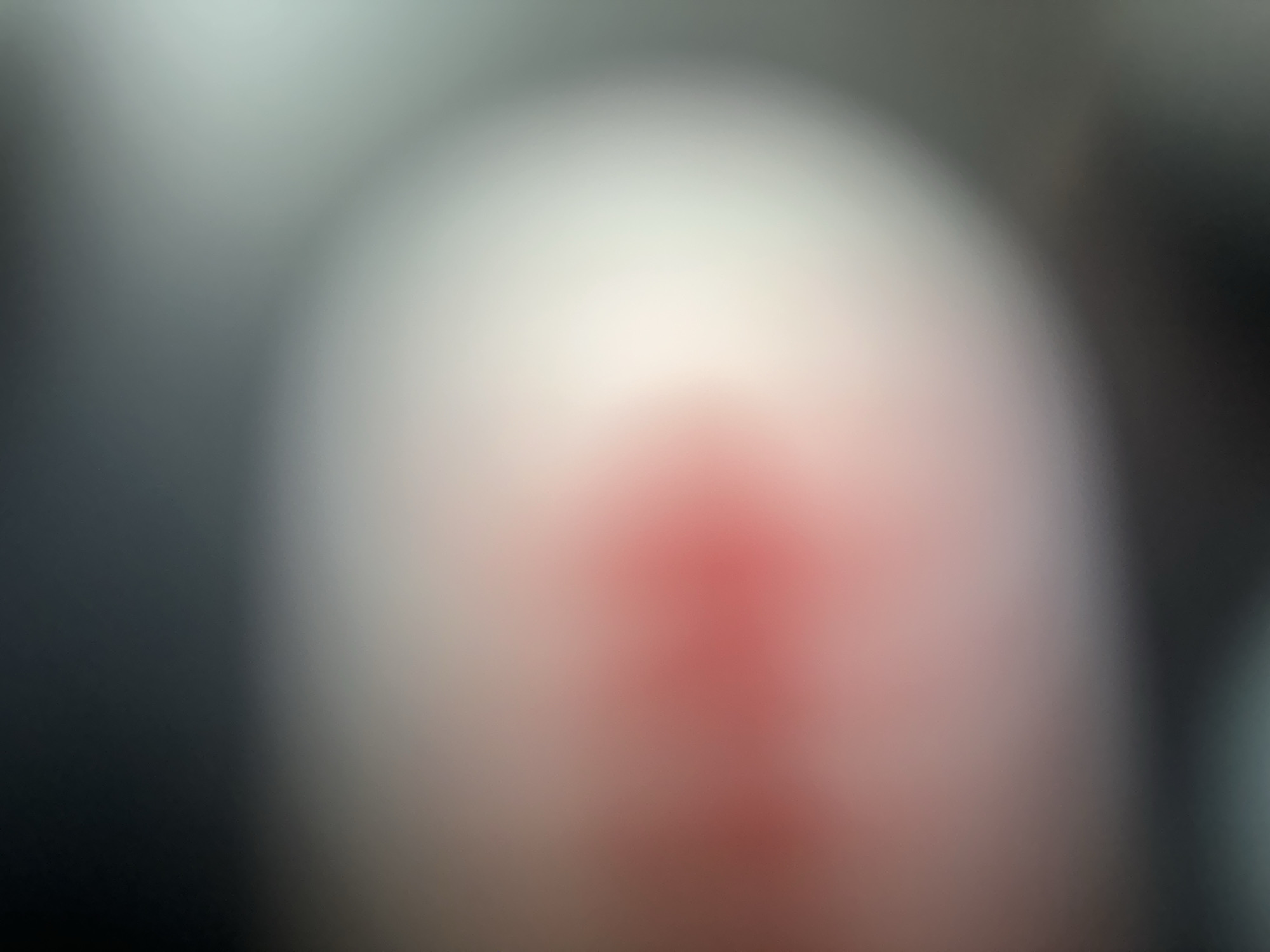 The image is an abstract, out-of-focus close-up of what appears to be a person's fingertip with a reddened area, possibly a wound or irritation, on its surface. The background is a blurry gradient of dark gray and lighter, muted tones, suggesting an indoor setting with soft lighting. The focus is entirely on the textured, slightly raised surface of the fingertip, with the reddish mark being the most prominent feature. There are no discernible objects, scene, setting, or activities beyond this close examination of skin. The lack of clear details makes it impossible to determine the time of day, weather, specific location within Belgrade, or any emotions. No text is visible.