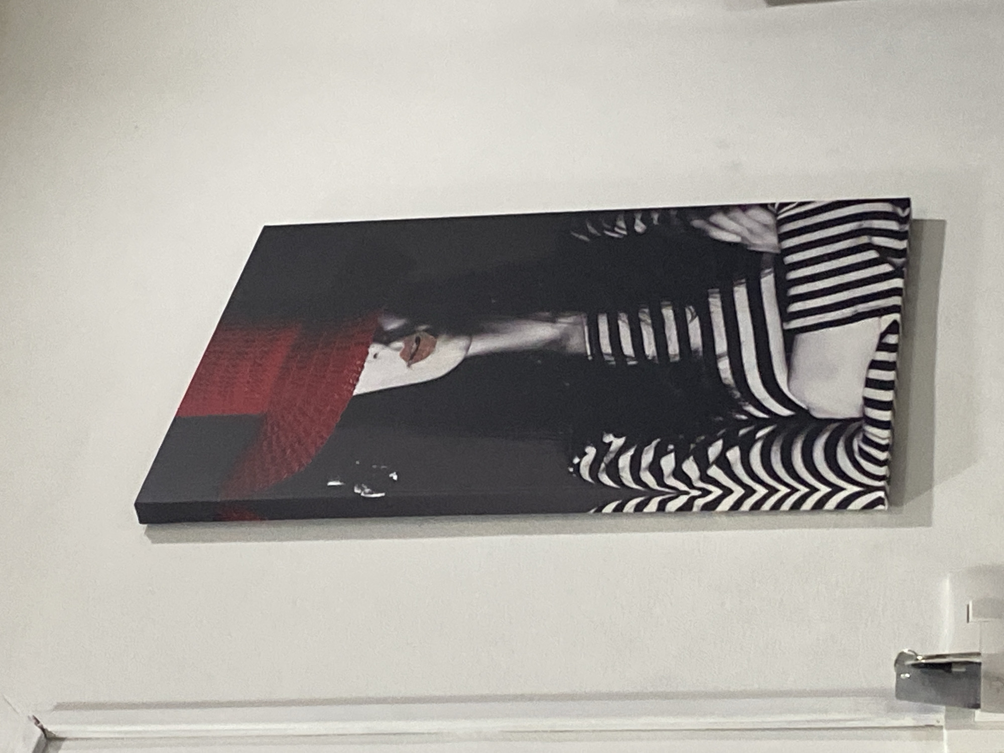 A horizontally oriented canvas print hangs on a plain, light-colored wall. The artwork on the print features a stylized portrait, dominated by black, white, and red tones, set against a dark background.
The central subject in the print is a person, likely a woman, depicted with a striking aesthetic. Their face is partially obscured by a wide-brimmed, textured red hat that covers the upper part of the face. Below the hat, prominent, bright red lips are visible. The subject is adorned in black and white striped clothing, which covers the body and limbs. The graphic stripes create a dynamic pattern across the garment, contributing to a high-contrast visual effect. The person appears to be posed at an angle, making the face seem sideways within the print's frame.
The wall is unadorned and shows a subtle shadow cast by the print, indicating its three-dimensional nature. At the very bottom of the overall image, a white trim or ledge is visible, along with a small metallic object in the bottom right corner, possibly a fixture. The setting appears to be indoors. There is no visible text in the image.