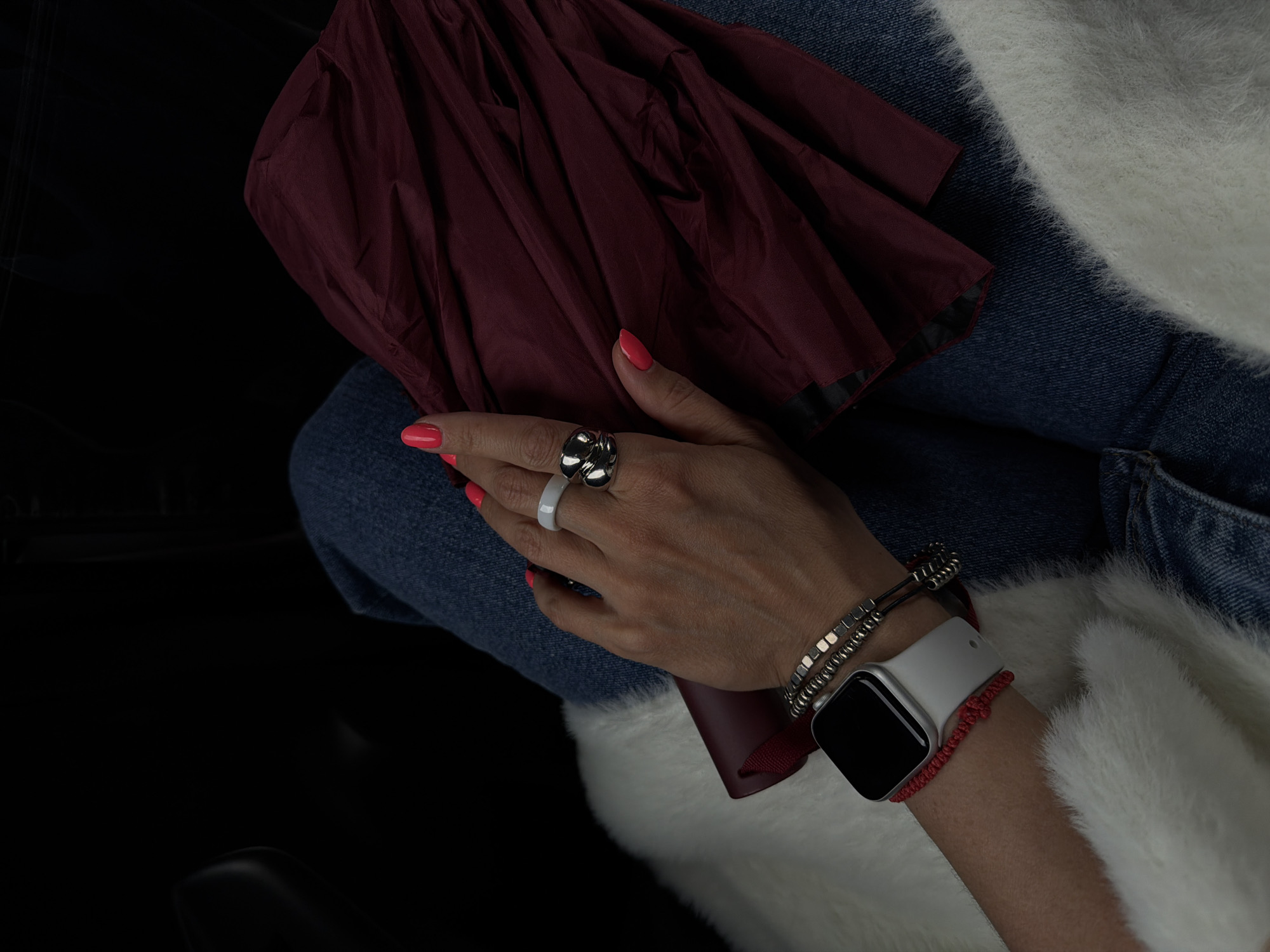 A woman's hand, adorned with a black floral ring and a white ceramic ring, is resting on a burgundy umbrella, which is in turn resting on her denim-clad lap. On her wrist, she wears a white smartwatch, a silver bracelet, and a red braided bracelet. She is wearing a white, furry jacket. The location appears to be indoors, possibly a car, given the dark background. The image contains no information indicating it was taken in the city of Birobidzhan, Russia.