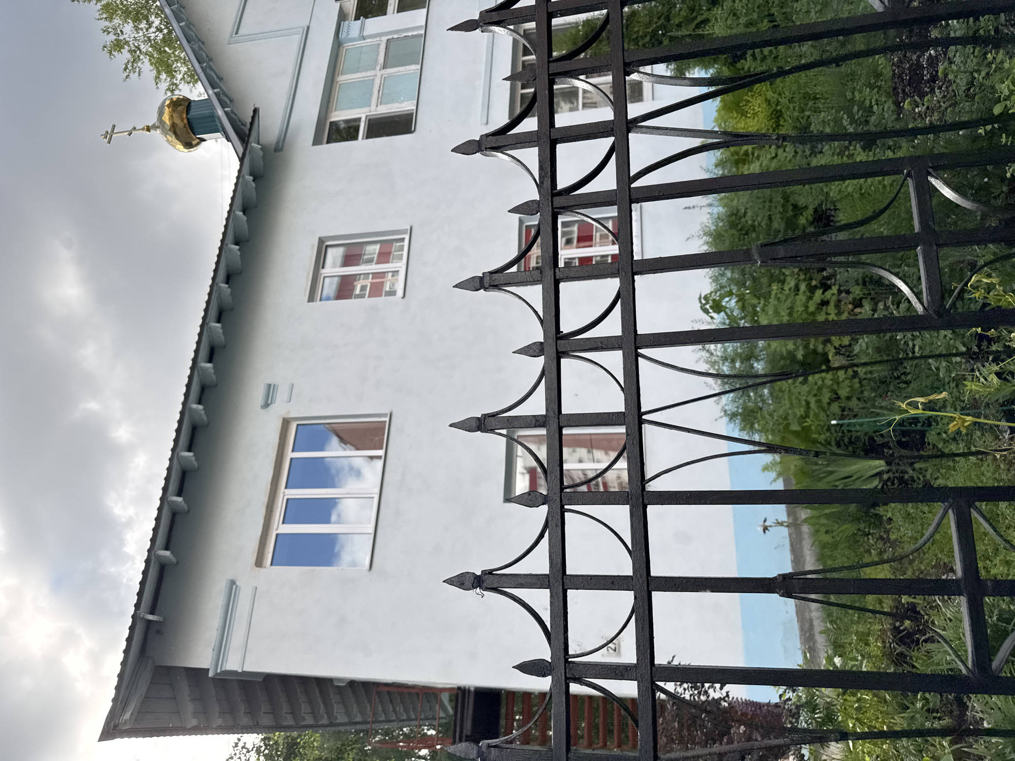 The image presents a ground-level, slightly upward-angled view of the Chapel of Saints Martha and Mary (Храм Святых Праведных Марфы и Марии) in Perm, Russia, located at Ulitsa Permskaya, 2A. A dark, ornate wrought-iron fence with pointed finials and decorative arches occupies the foreground, partially obscuring the lower section of the building and the garden area.
The chapel is a two-story structure with light blue-grey stucco walls and a dark grey corrugated roof. The roofline features decorative light blue brackets supporting the eaves. On the right side of the building's roof, a small, dark green drum supports a golden, onion-shaped dome topped with a golden Orthodox cross, glinting under the cloudy sky.
The building features multiple windows with white frames. The upper left window reflects a cloudy sky with hints of a light blue horizon and a distant, reddish building. Other windows, particularly those visible through the fence, also show reflections of the surrounding environment, including a prominent red-colored structure or wall. A small, decorative light blue architectural element is present below the roofline on the upper left section of the building. On the left side, a dark, possibly wooden, gable roof section extends downwards, and a red external staircase or railing is partially visible behind the building.
Below the chapel's walls and visible through the fence, a lush, overgrown garden or yard contains various green plants, shrubs, and some taller, slender plants. The sky above is overcast with textured grey and white clouds. No people are visible in the scene.