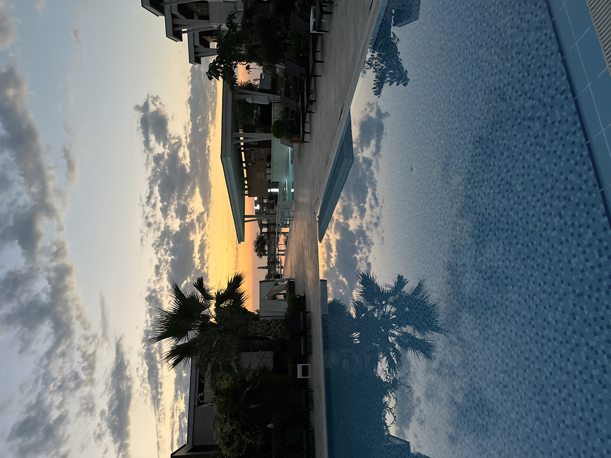 The image depicts a tranquil resort scene at dusk in Sozopol, Bulgaria, with a focus on a swimming pool reflecting the sunset and surrounding architecture. In the foreground, a large swimming pool with blue tiled bottom dominates the lower half of the frame. The water's surface is calm, acting as a mirror for the sky and palm trees.
To the left and center of the mid-ground, several dark, modern sun loungers are arranged on a tiled patio. A large palm tree stands prominently to the left, its fronds casting a distinct reflection on the pool. Behind the loungers and palm tree, a white cabana structure with sheer curtains is visible.
Further into the mid-ground, a modern, open-air bar structure with a white roof is present. The bar area is illuminated, and signs indicating "POOL BAR" and "Gelateria" are discernible. Beyond the bar, a walkway leads towards more distant resort buildings and a hint of the sea, bathed in the warm hues of the setting sun.
The sky above is a canvas of twilight colors, with streaks of orange and yellow from the setting sun transitioning into soft blues and grays. Wispy clouds are scattered across the sky, adding texture and depth to the scene. The architecture visible is contemporary, featuring clean lines and geometric shapes, consistent with a modern resort design. The overall atmosphere is peaceful and relaxing, suggesting the end of a warm day.