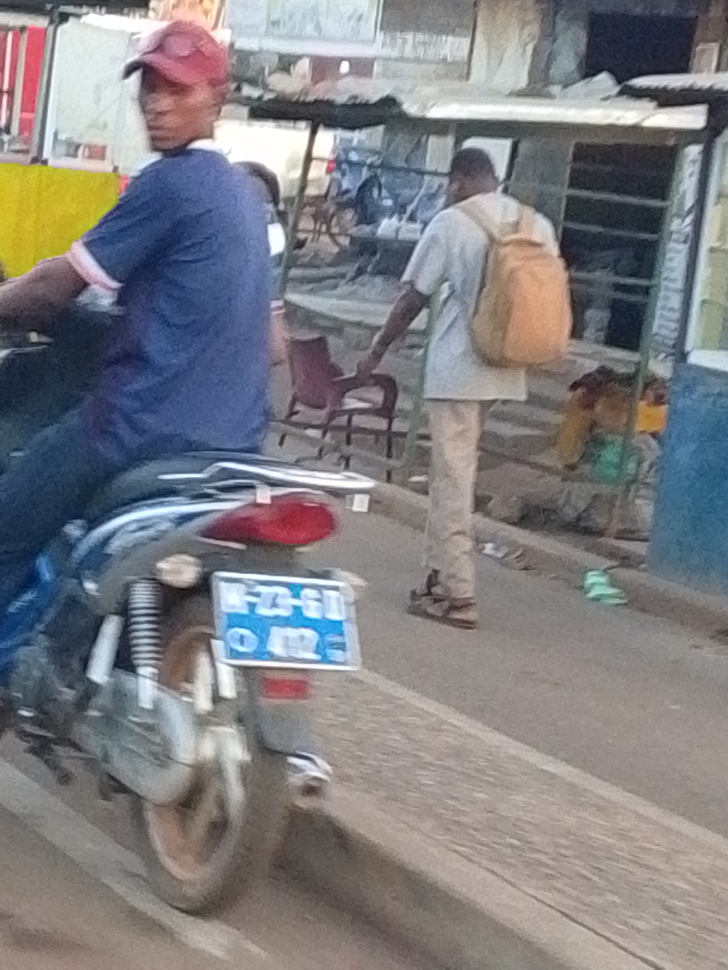 The image depicts a street scene in Tamale, Ghana. A man wearing a red cap and a blue shirt is sitting on a parked moped with a blue license plate reading "N-23-GI 012." Another man, wearing light-colored pants and shirt with a tan backpack, is walking away from the camera on the sidewalk. In the background are various buildings, including what appears to be small shops or stalls. A red chair can be seen along the road and some bikes in the background. The weather appears to be dry and sunny.