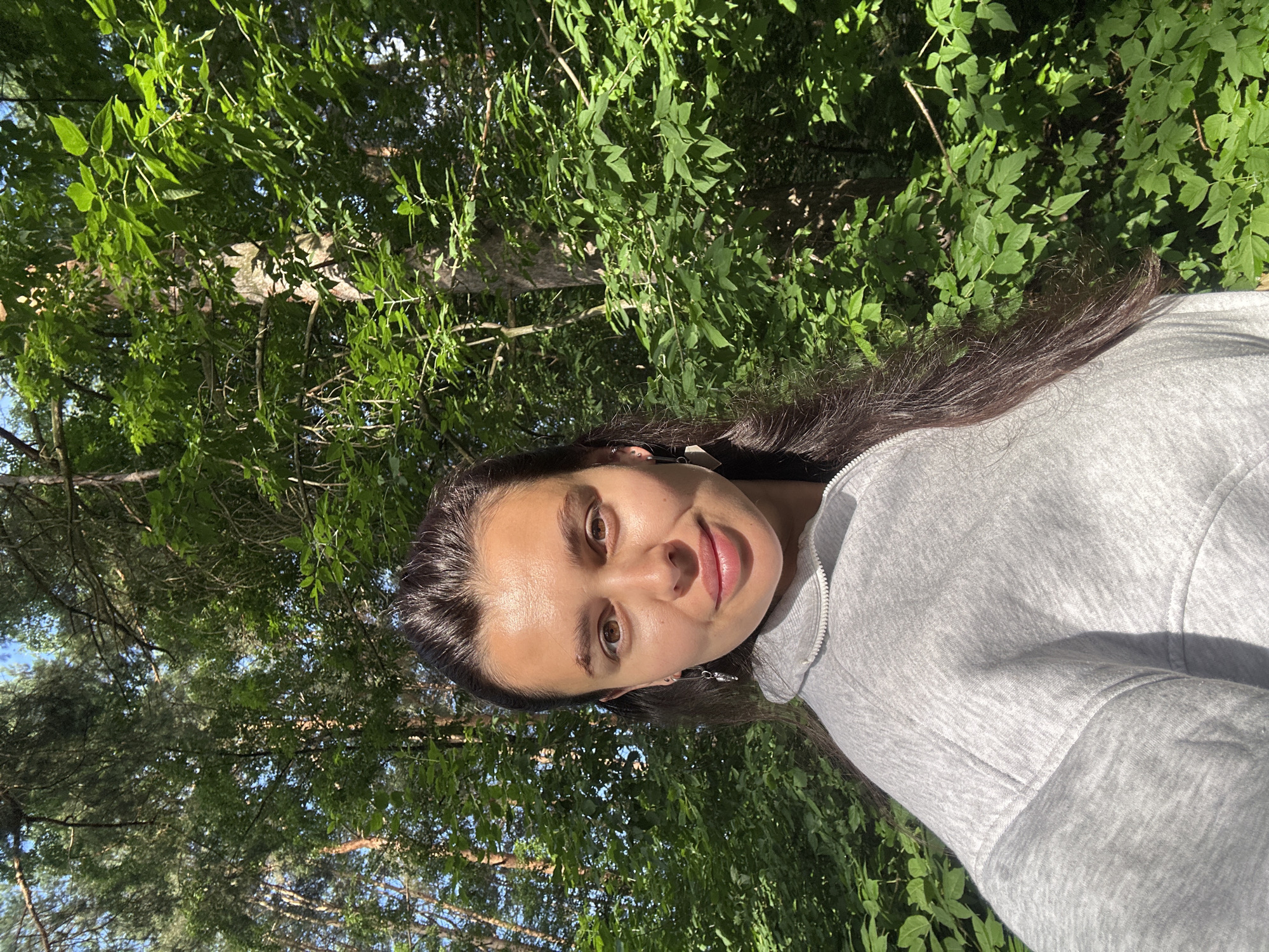 The image shows a young woman with long, dark hair and brown eyes, wearing a grey zip-up hoodie. She is positioned in the foreground, facing the camera with a slight smile. Behind her, lush green foliage and trees suggest an outdoor, natural environment. The lighting indicates it is daytime, with sunlight filtering through the leaves. The specific location is a forest area, likely on the outskirts or within a park in Chaikovsky, Russia, given the types of trees and the overall atmosphere.