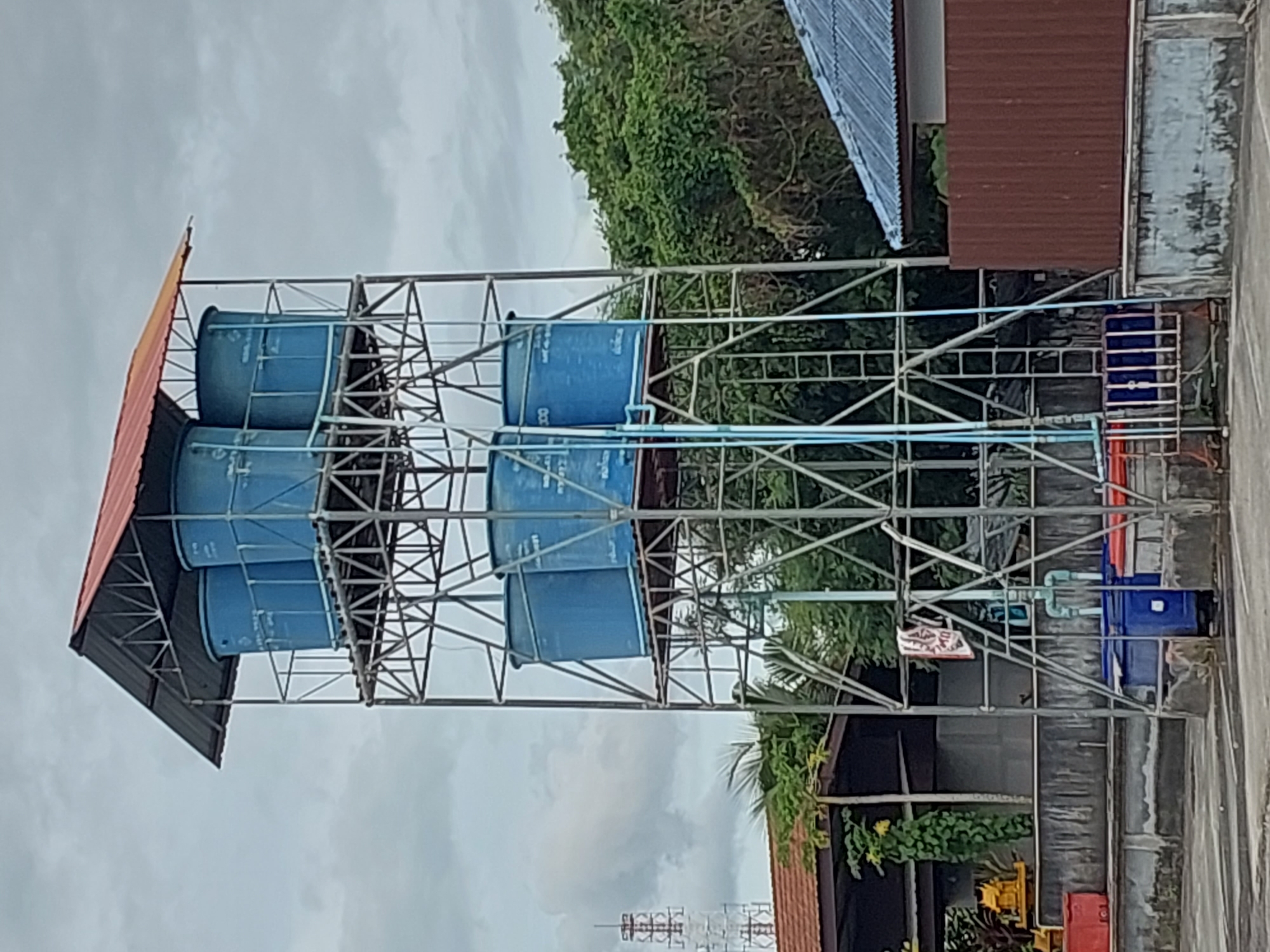 The image shows a water tower in Baan Mae Nam, Thailand, under a cloudy sky. The water tower is a tall, metallic structure supporting several large blue cylindrical water tanks on multiple platforms. The uppermost platform is covered by a red, angled roof supported by metal beams. Below the tanks, a ladder is attached to the tower's frame, and blue pipes run down the side. At the base of the tower, two large blue containers are visible, along with some red containers to the left. In the background, a tall communication tower can be seen in the distance, along with lush green trees and buildings with red and blue roofs. The scene appears to be an outdoor industrial or utility area.