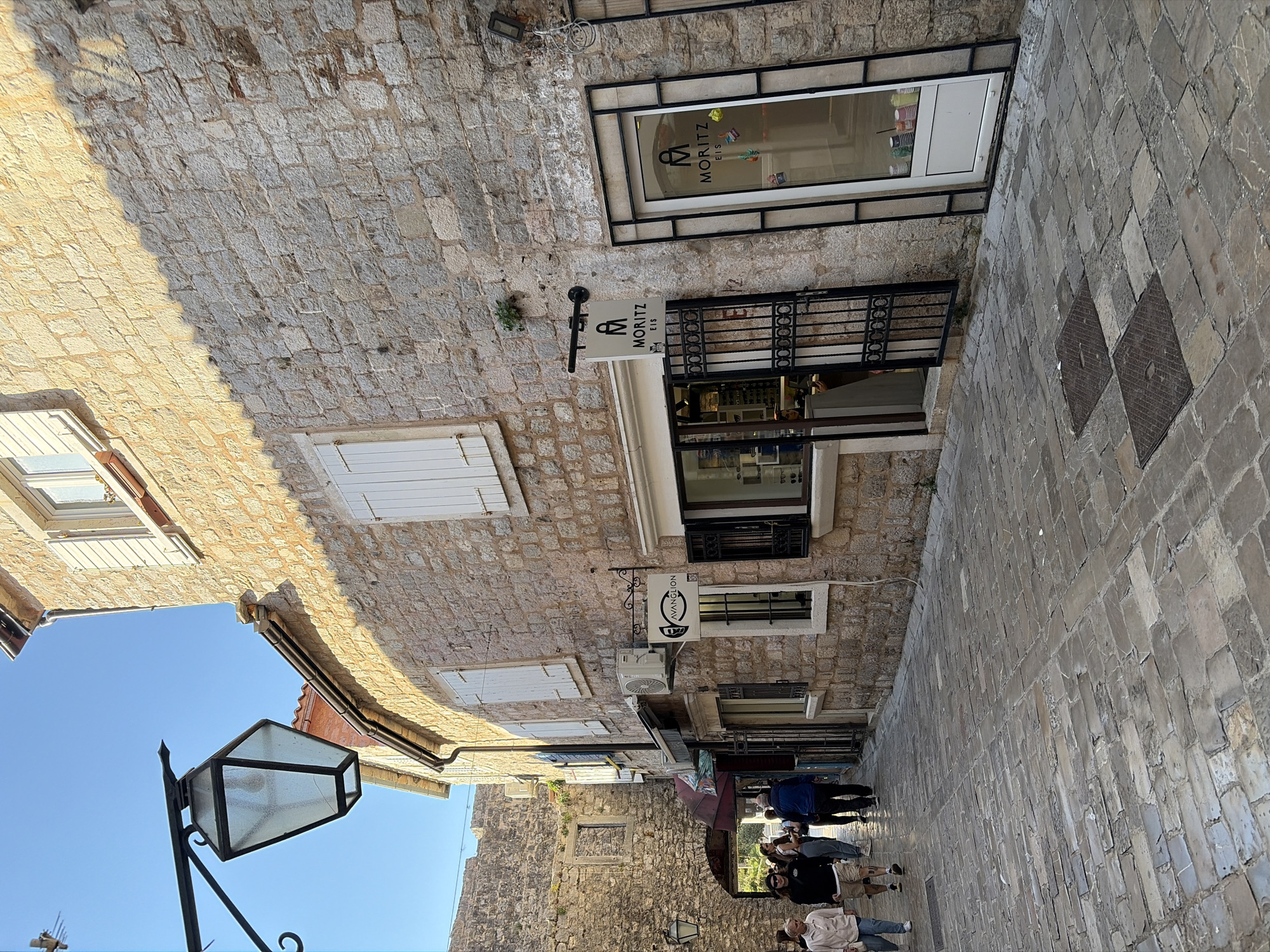 The image depicts a narrow street in the old town of Budva, Montenegro. The street is paved with light gray stones. The buildings on either side are constructed from light-colored stone blocks. Many of the buildings have windows with white shutters and black metal grates. There is a store called "Moritz Eis" (ice cream) on one side of the street. People are walking along the street, including a group of people and individuals dressed in casual clothes. There is a black lamppost on the left side of the street. The sky is blue in the top left corner of the image.