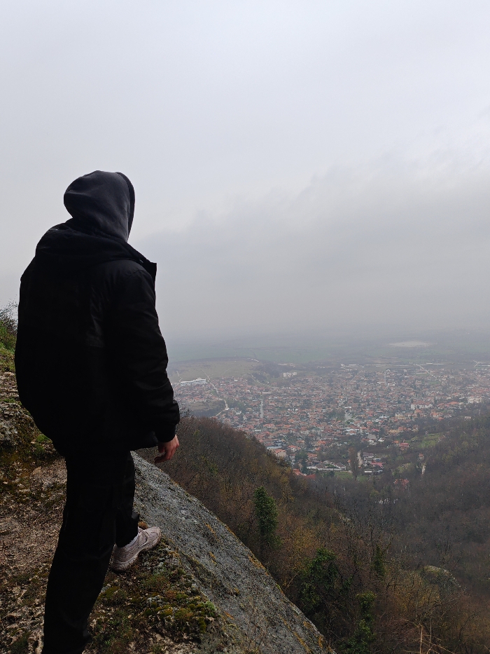 A solitary individual stands with their back to the viewer on a moss-covered rock ledge, positioned at an elevated viewpoint overlooking the city of Shumen, Bulgaria. The person wears a black hooded jacket with the hood up, black trousers, and light-colored athletic shoes. The individual is oriented towards the urban landscape below, which comprises a dense arrangement of buildings, many featuring reddish-brown roofs, bordered by forested hillsides and agricultural fields in the distance. The immediate foreground shows the textured grey rock of the ledge sloping sharply downwards. The environment is under an uniformly overcast, grey sky, with atmospheric haze obscuring the distant horizon. The location is consistent with a viewpoint on the Shumen Plateau overlooking the city.