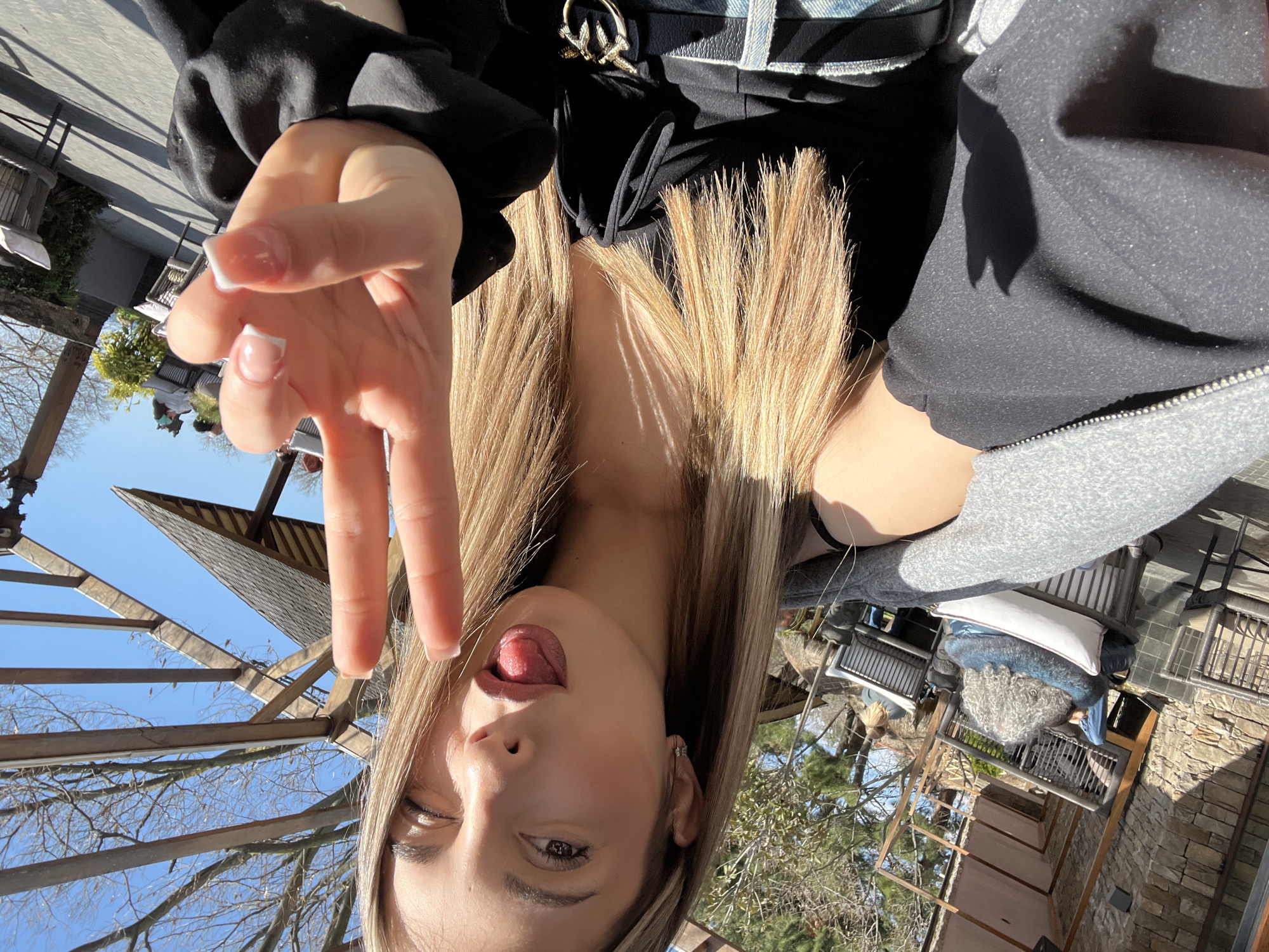 A young woman is taking a selfie, tilting her head to the side and sticking out her tongue. She is making a peace sign with her fingers. She has blonde hair and wears black clothing. The background suggests an outdoor seating area, possibly a cafe or restaurant. The furniture consists of a grey chair with a white pillow, a person sitting in it, and a wrought iron fence. The sky is blue. The exact location in Varna, Bulgaria, cannot be determined from the image.