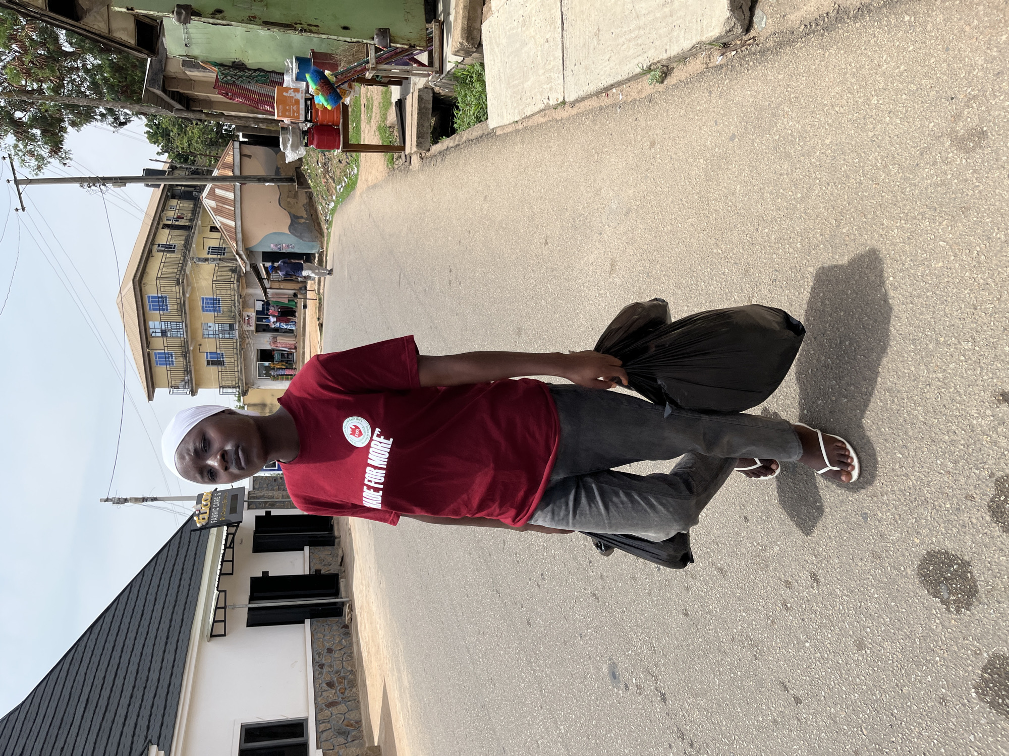 A young man stands on a paved road in Jos, Nigeria. He wears a maroon t-shirt with white lettering, grey pants, and white sandals. He also wears a white cap. He holds two black plastic bags. A two-story building with a yellow facade and blue windows stands in the background. Across the road, there's a white building with black-framed windows and a dark grey roof. Beside it, there's a green metallic structure. The sky is overcast.