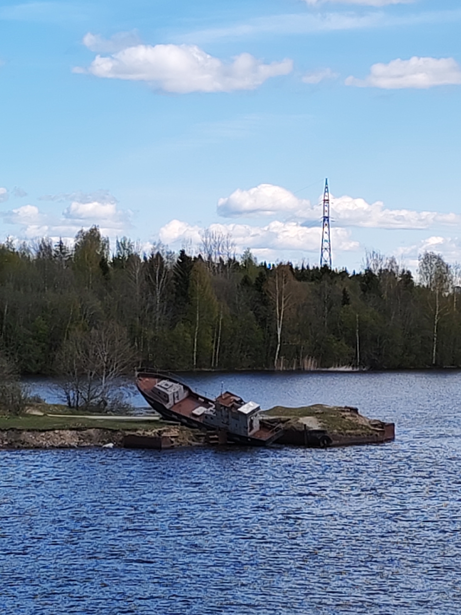 The image shows an old, possibly wrecked, vessel resting on a small, grassy island in a body of water. The vessel appears to be partially submerged or tilted significantly. The surrounding water is choppy with a blue hue. In the background, a forested area is visible with trees of varying green shades. A tall, thin, blue-and-red lattice tower rises above the treeline. The sky is blue with scattered white clouds. The location is Ivanov Bor, Russia.