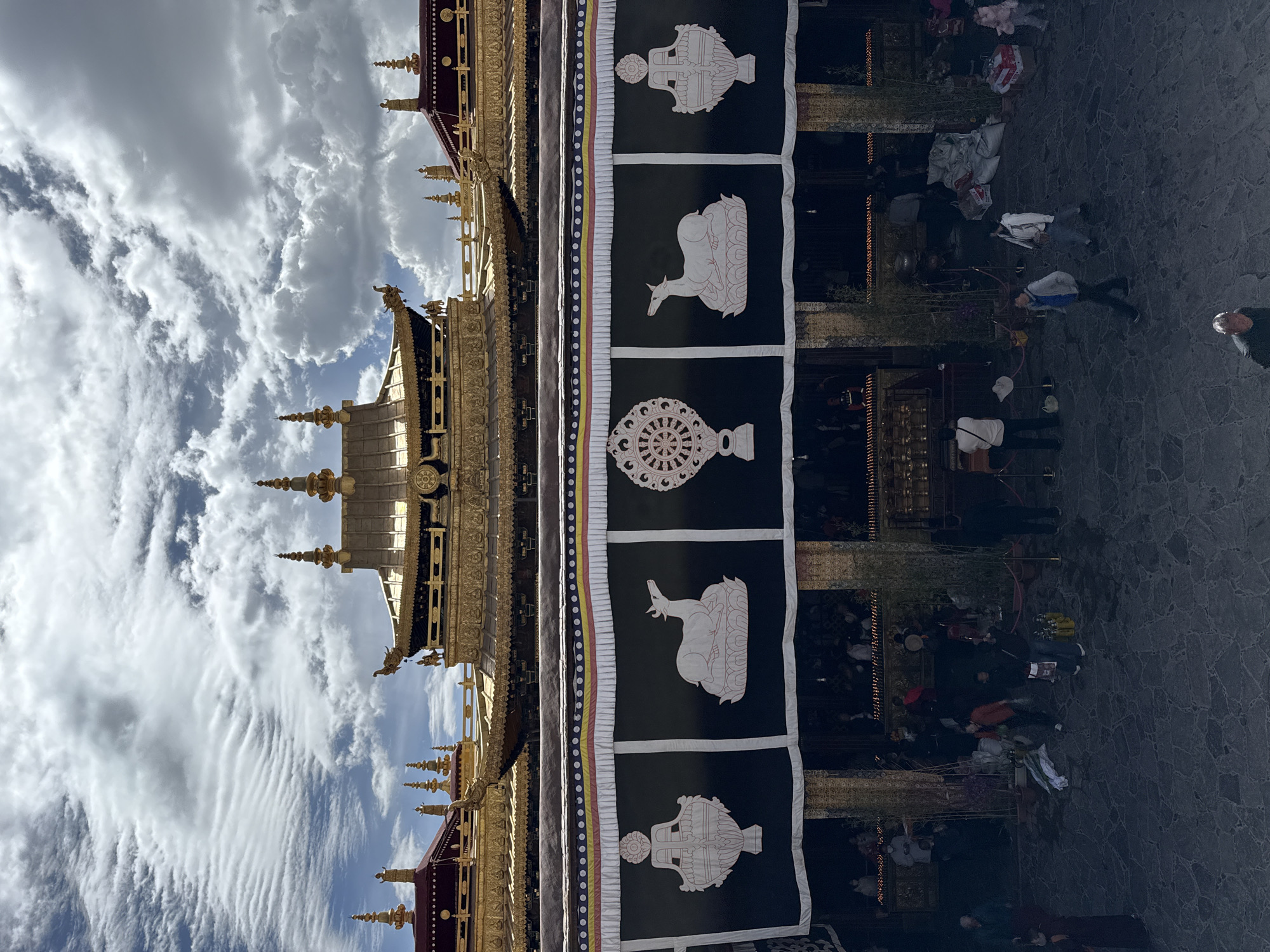 The image shows a low-angle view of a building with golden ornate structures against a cloudy sky. A black banner with white symbols and white lines is hanging from the building. These symbols, from top to bottom, depict a treasure vase, a pair of deer, and a dharma wheel. The bottom part of the image shows a courtyard with stone paving where people are walking or standing. The location is likely in the Chengguan District, China, given the style of architecture.