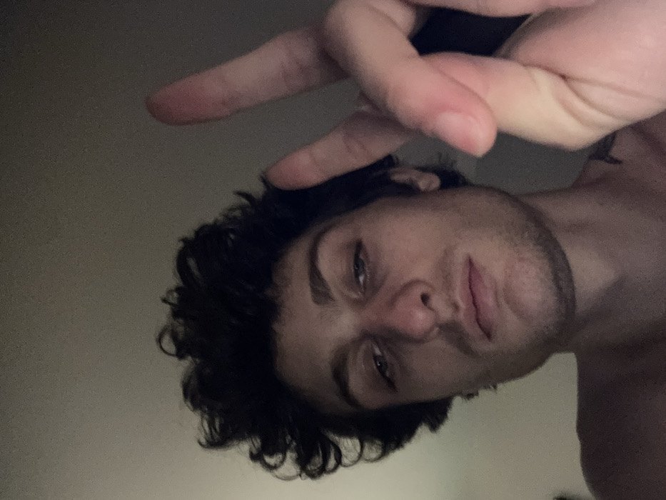 A close-up, low-angle selfie depicts a male subject in an indeterminate indoor setting in the city of Vincennes, France. The male, appearing to be in his 20s-30s, has dark, curly hair and a light complexion. His face is centrally framed, featuring a prominent nose with some redness on the tip and bridge, closed lips, and dark, heavy-lidded eyes looking directly forward. Slight facial stubble is visible on his jawline and chin. His bare shoulders and upper chest are visible, with a faint dark mark on his upper right shoulder/neck area. His right hand is raised to the right of his head, with the index and middle fingers extended in a peace sign gesture. The background consists of a plain, uniformly light-colored wall or ceiling, dimly lit.