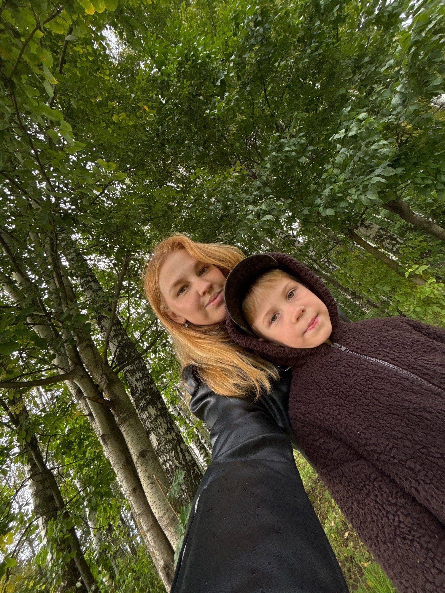 An outdoor, low-angle, medium shot shows a woman with long blonde hair on the left and a young boy wearing a dark brown hooded jacket on the right, standing close together amidst a grove of trees. The woman is wearing a black, possibly leather, jacket. The boy's face is partially obscured by his hood. The trees are tall with white and dark bark, and green foliage. The ground beneath is covered in grass and some fallen leaves. The image appears to be taken in a park or forest, likely within the city of Yaroslavl, Russia, as suggested by the context.