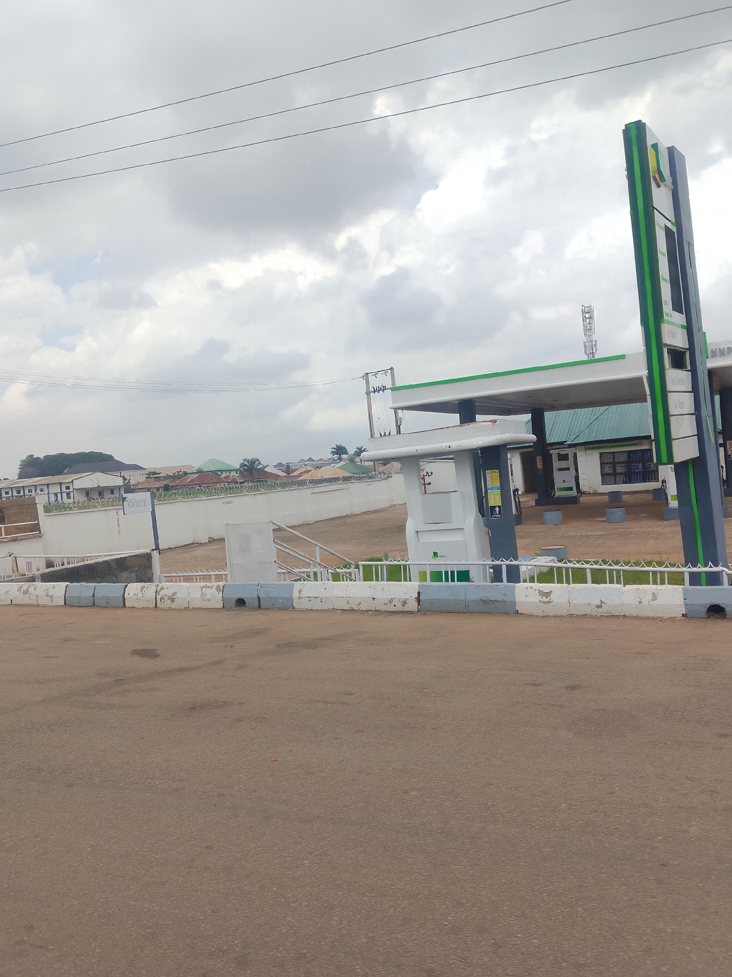 An overcast, medium-shot, eye-level, outdoor, daytime, wide-angle image shows a gas station with a towering, green and white sign. Three power lines stretch across the top of the frame. A white concrete barrier with blue and grey painted stripes runs across the bottom. The gas station has a white and green canopy, with multiple fuel pumps visible underneath. Behind the gas station, a row of buildings with red and green roofs is visible. A tall, cellular tower stands in the background. The ground in front of the gas station is covered with sand and gravel. The area appears to be a main road with a paved surface.