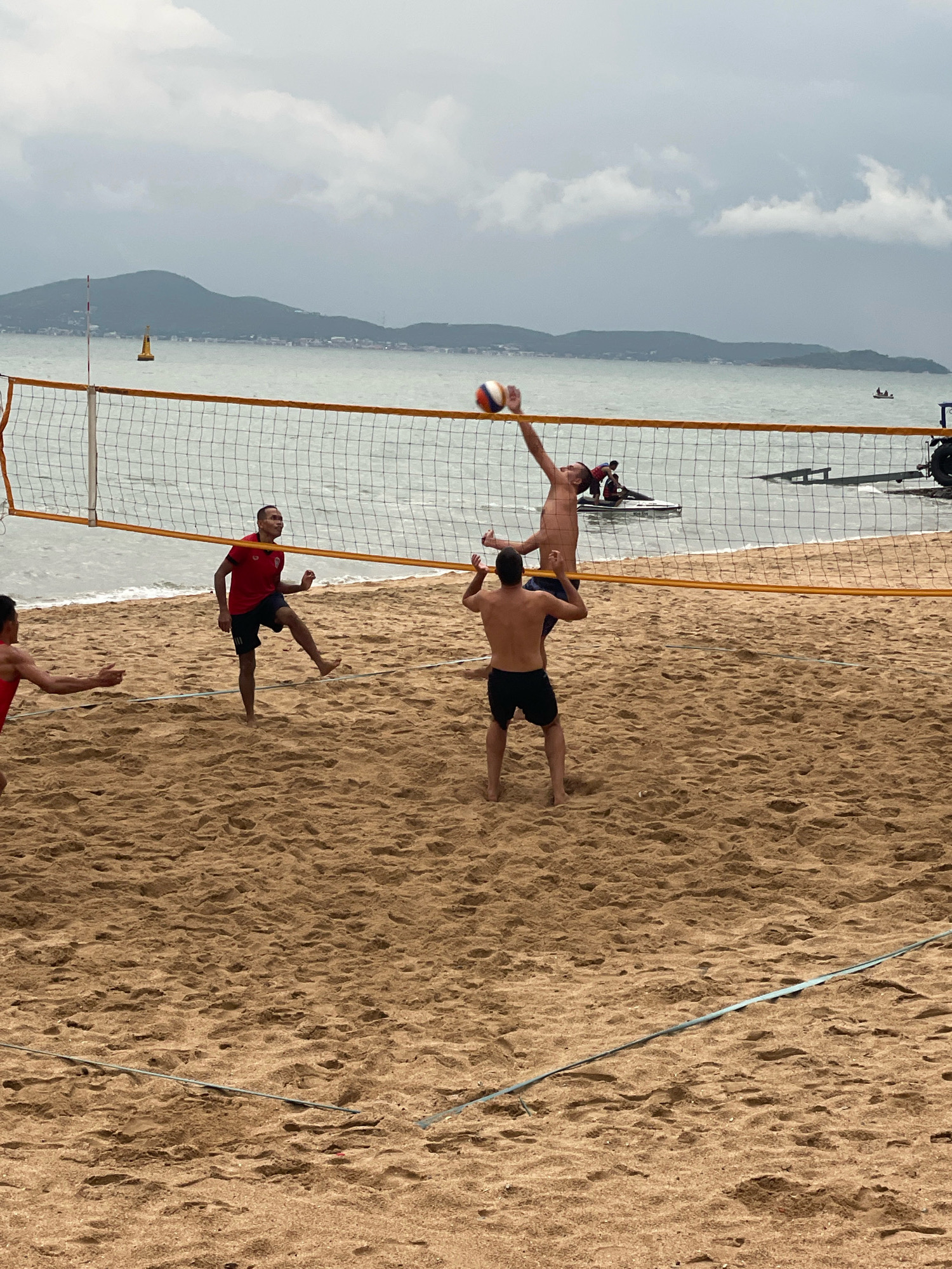 Four men are playing beach volleyball on a sandy beach in Pattaya City, Thailand. A volleyball net is set up across the sand, with the ocean and a cloudy sky visible in the background. One man in a red shirt and black shorts is on the left side of the net, kicking the ball up with his right foot. On the right side of the net, two men are in mid-action. One man, shirtless, is jumping to spike the ball, while another man, also shirtless, is positioned to receive the ball. The fourth man, shirtless, is watching the play unfold. The beach is a light brown sand, with footprints and some debris visible. In the background, a few boats are on the water, and a hilly coastline can be seen. The weather appears overcast, suggesting a potentially stormy day.