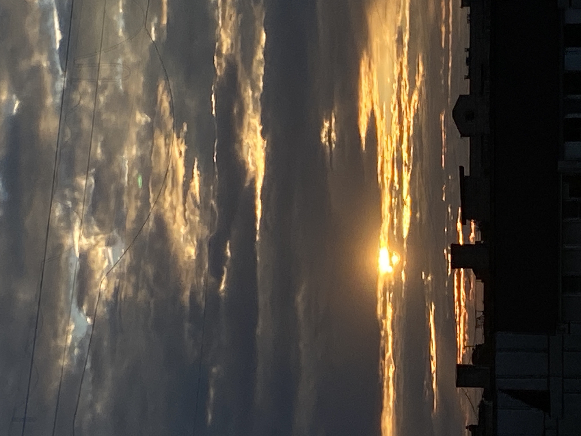 A dramatic sunset illuminates a cloudy sky over the city of Chaikovsky, Russia. Dark, textured clouds are lit from behind by the sun, creating streaks of golden light and casting deep shadows. The sun itself is a bright, diffused orb peeking through a break in the clouds. Below the sky, the silhouettes of buildings form a dark, urban skyline. The rooftops are varied, with some appearing flat and modern, while others have pitched roofs and chimneys. Power lines are visible crisscrossing the upper portion of the image, adding to the urban feel. The overall impression is one of a moody, atmospheric evening.