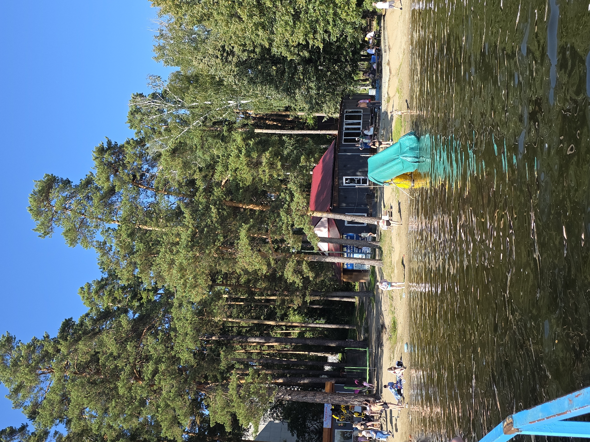 The image shows a sunny day at a beach in Chebarkul, Russia. On the sandy shore, numerous people are visible, some sunbathing, others walking or standing. A green water slide with a yellow base is set up near the water's edge, and a few people are interacting around it. Several buildings are situated behind the beach, one with a red roof and dark walls, appearing to be a cafe or service area. Tall pine trees and other deciduous trees form a dense forest backdrop. The foreground is dominated by the dark, rippling surface of the water, reflecting the surrounding greenery and sky. A blue railing of a boat or pier is partially visible in the bottom left corner.